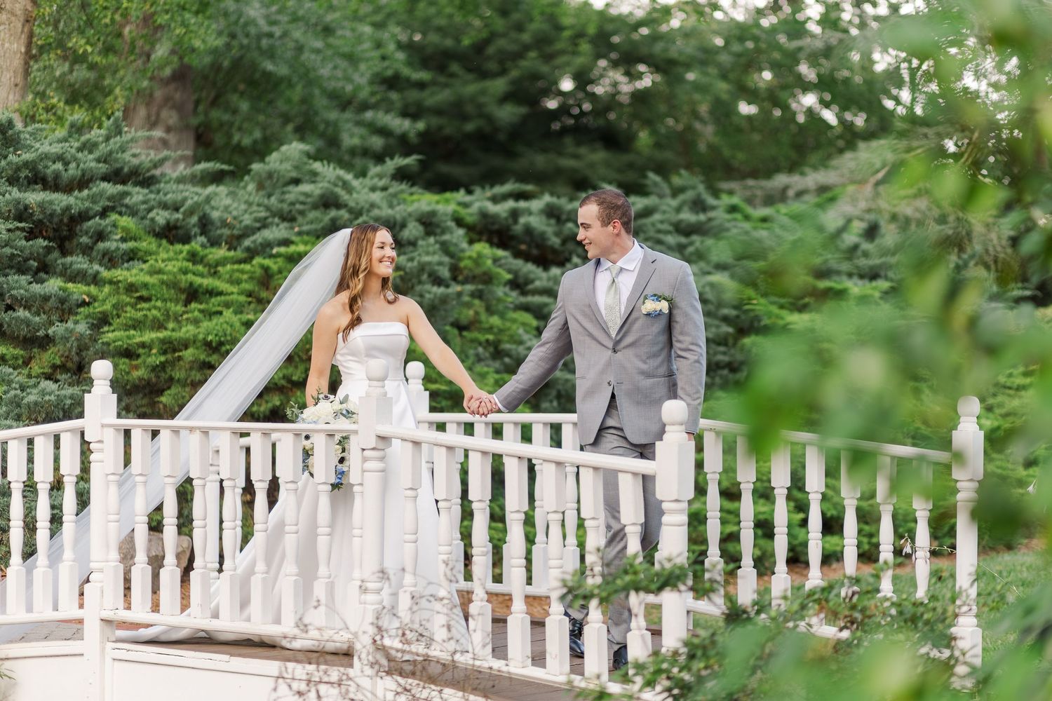 Wedding couple poses on white balustrade walkway surrounded by lush greenery.