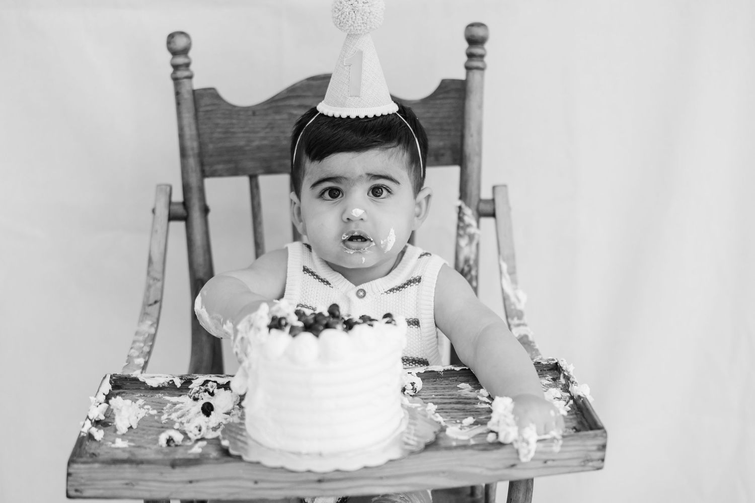 Black and white photo of a baby in a high chair wearing a party hat with a birthday cake, celebrating their first birthday.