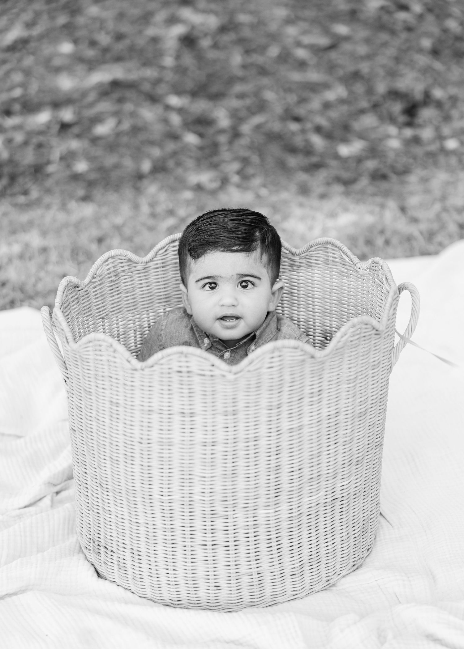 Black and white photo of a cute baby sitting inside a large wicker basket outdoors on a white blanket.