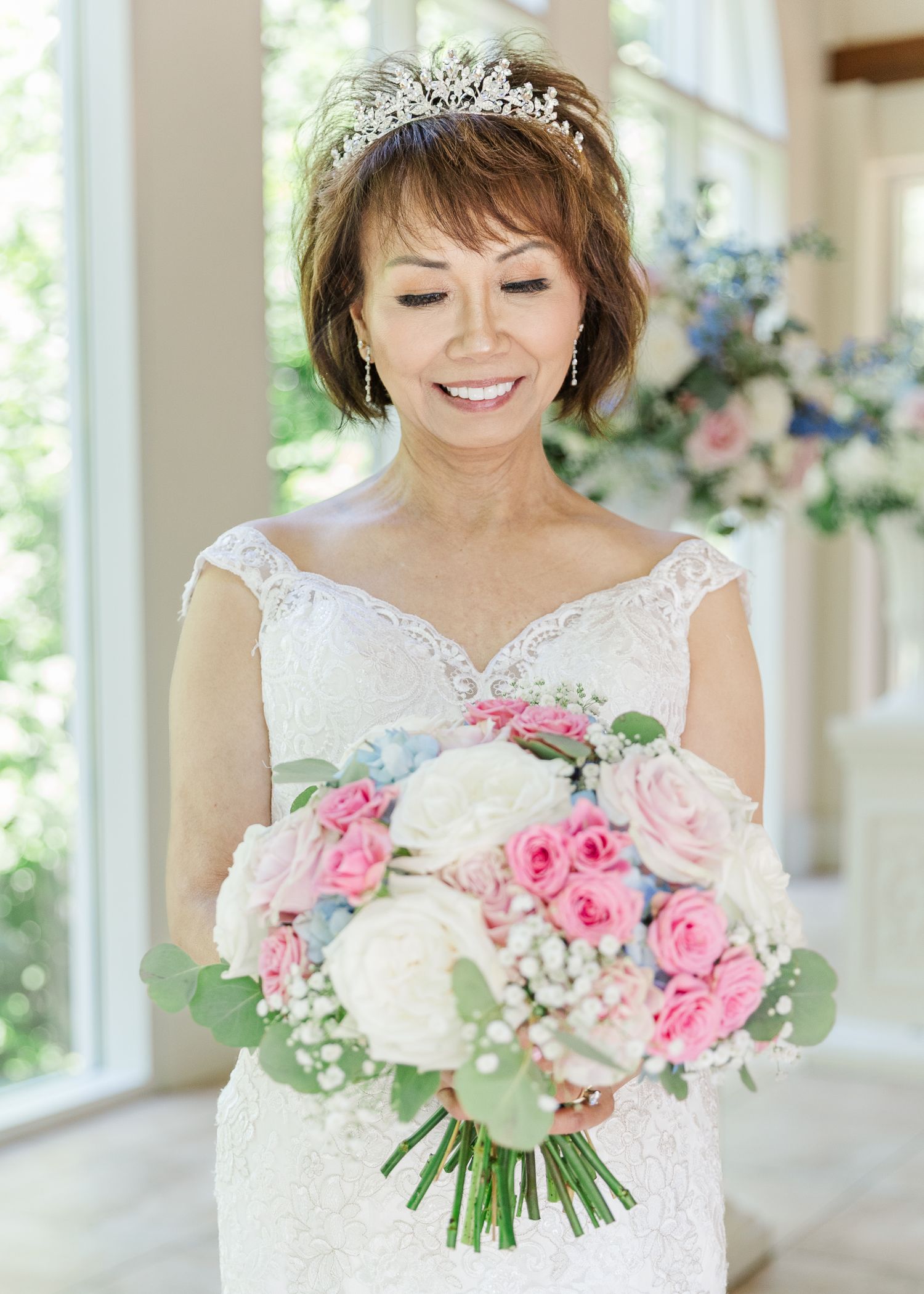 Smiling bride in lace wedding dress and tiara holds pink and white rose bouquet indoors.