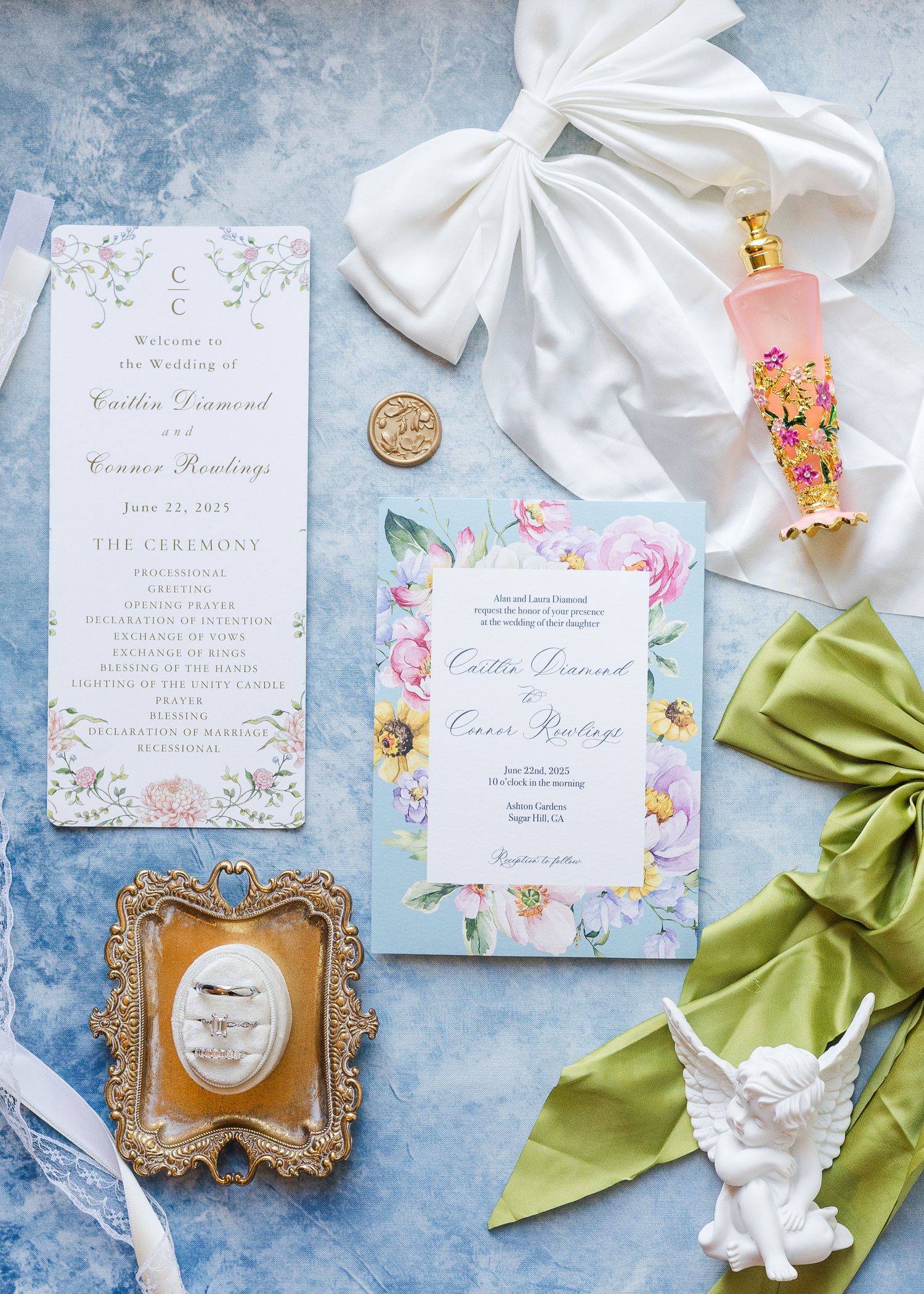 Elegant wedding stationery flat lay with floral invitations, perfume bottle, angel figurine, and silk ribbons on blue backdrop.