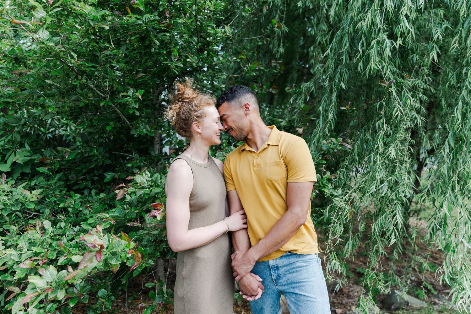 A couple smiles at each other while holding hands among lush green trees outdoors.