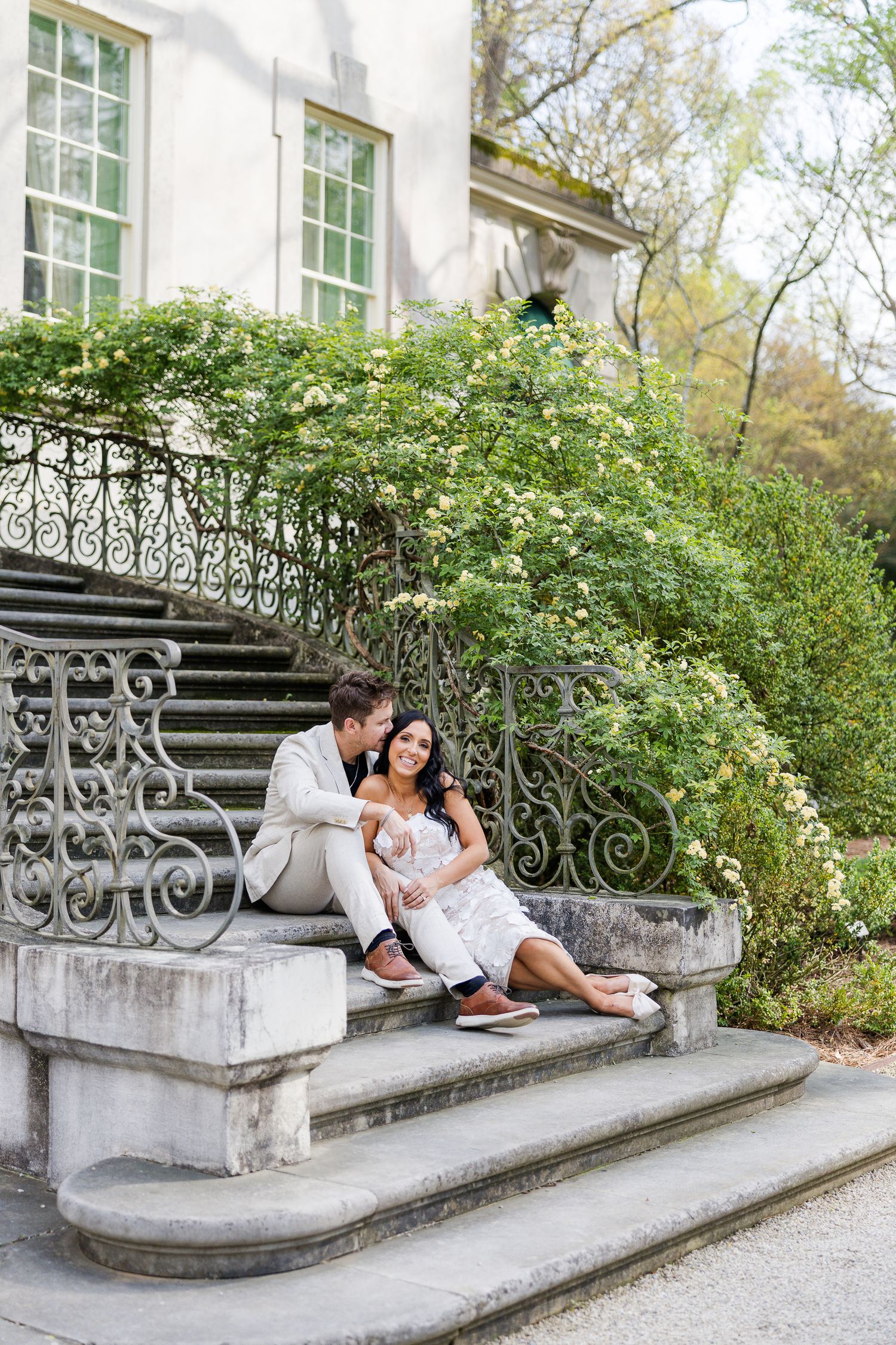 Couple sitting on ornate stone steps of elegant mansion, man in tan suit kissing woman in white dress outdoors.