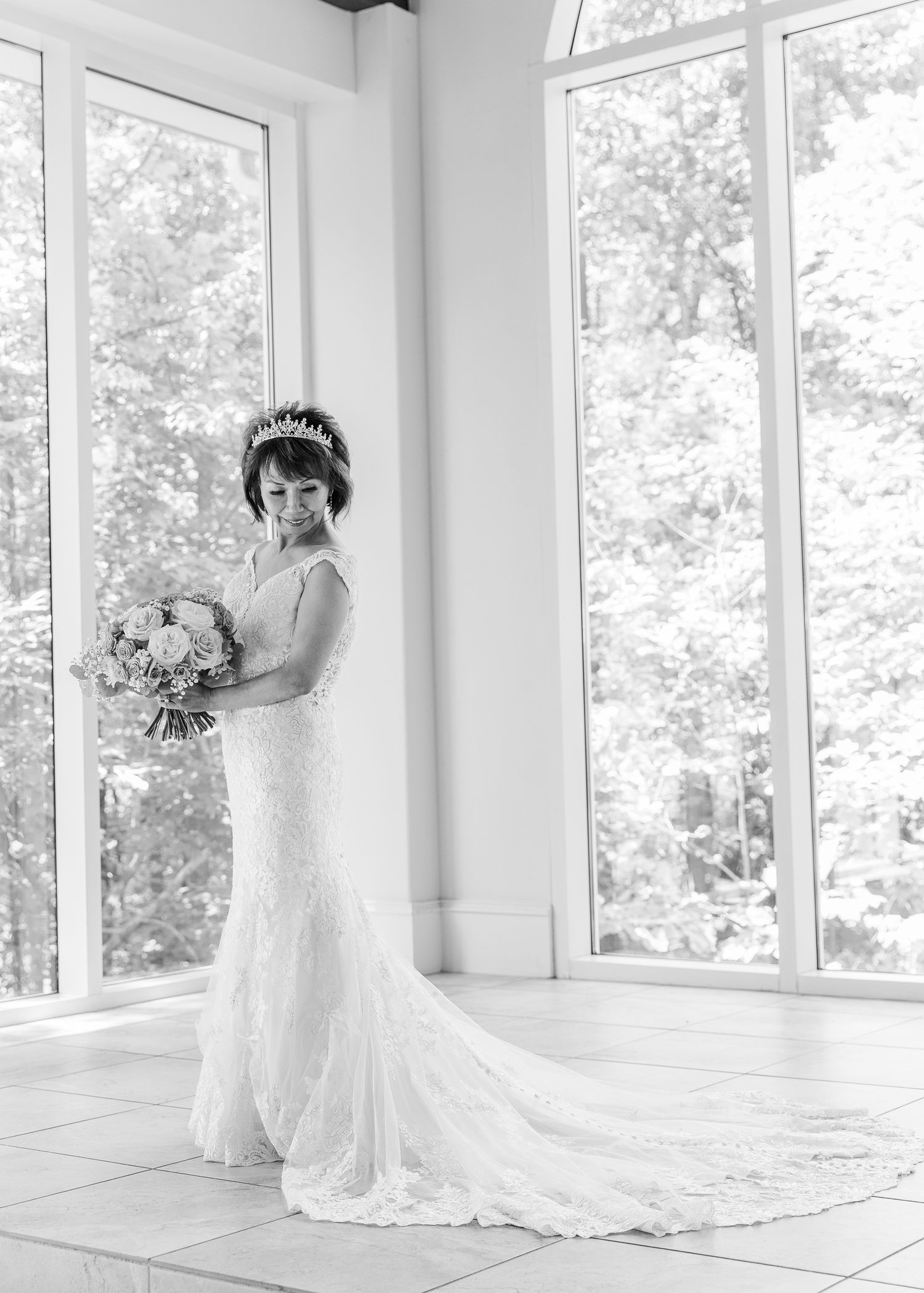 Bride in lace mermaid gown and tiara holds bouquet by large windows in elegant black and white bridal portrait.