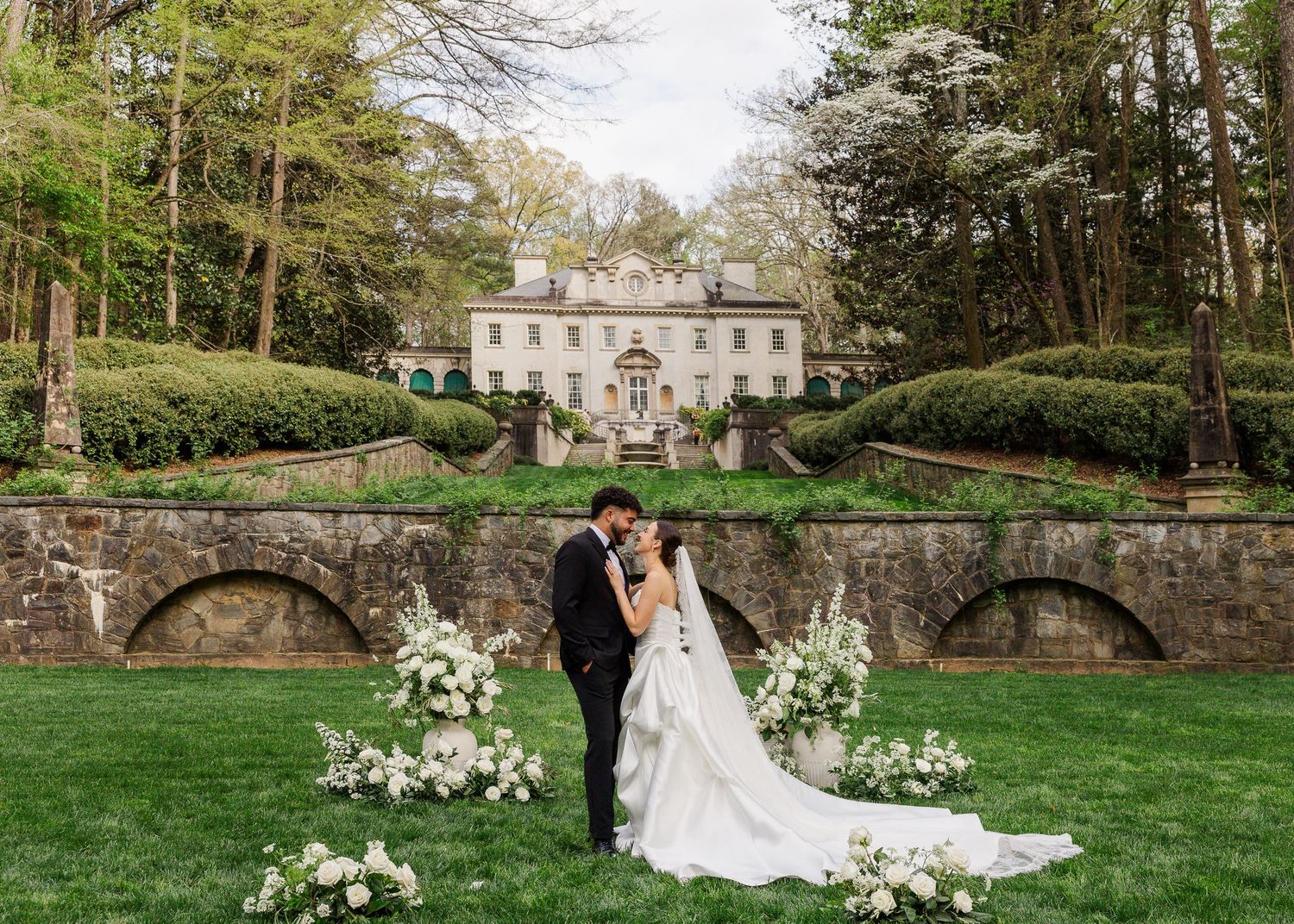 Bride and groom share a romantic kiss in front of an elegant estate with lush gardens and floral arrangements.