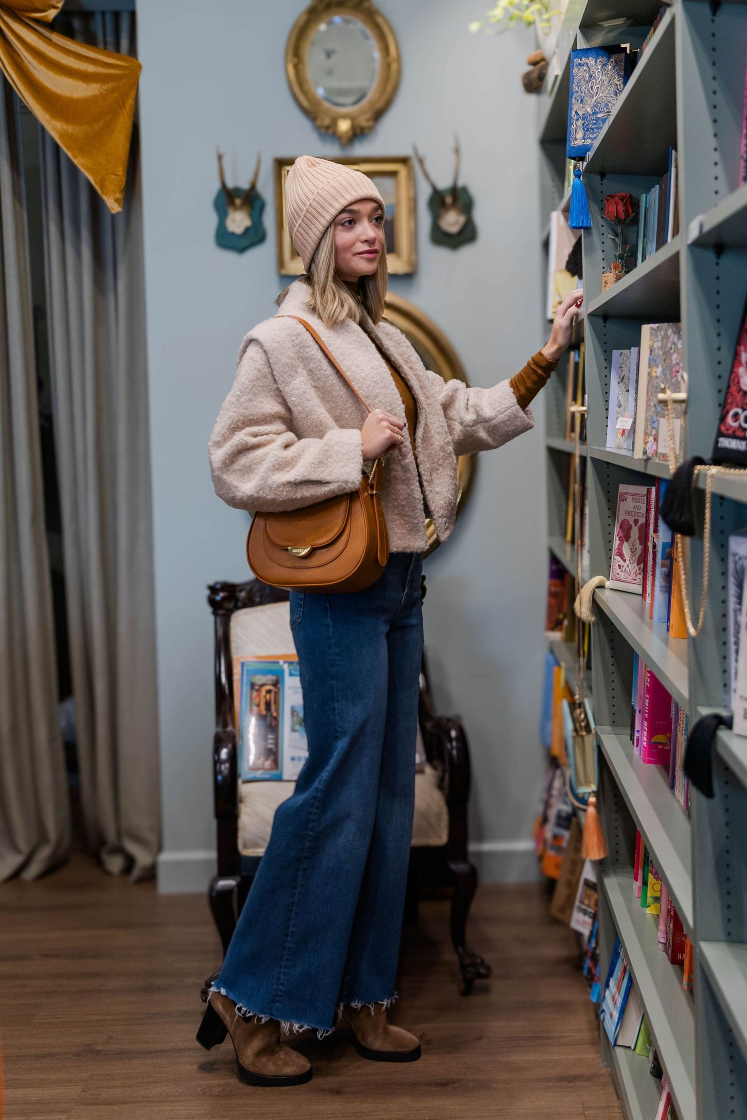 Beige coat and wide-leg jeans styled with brown bag while browsing bookstore shelves.