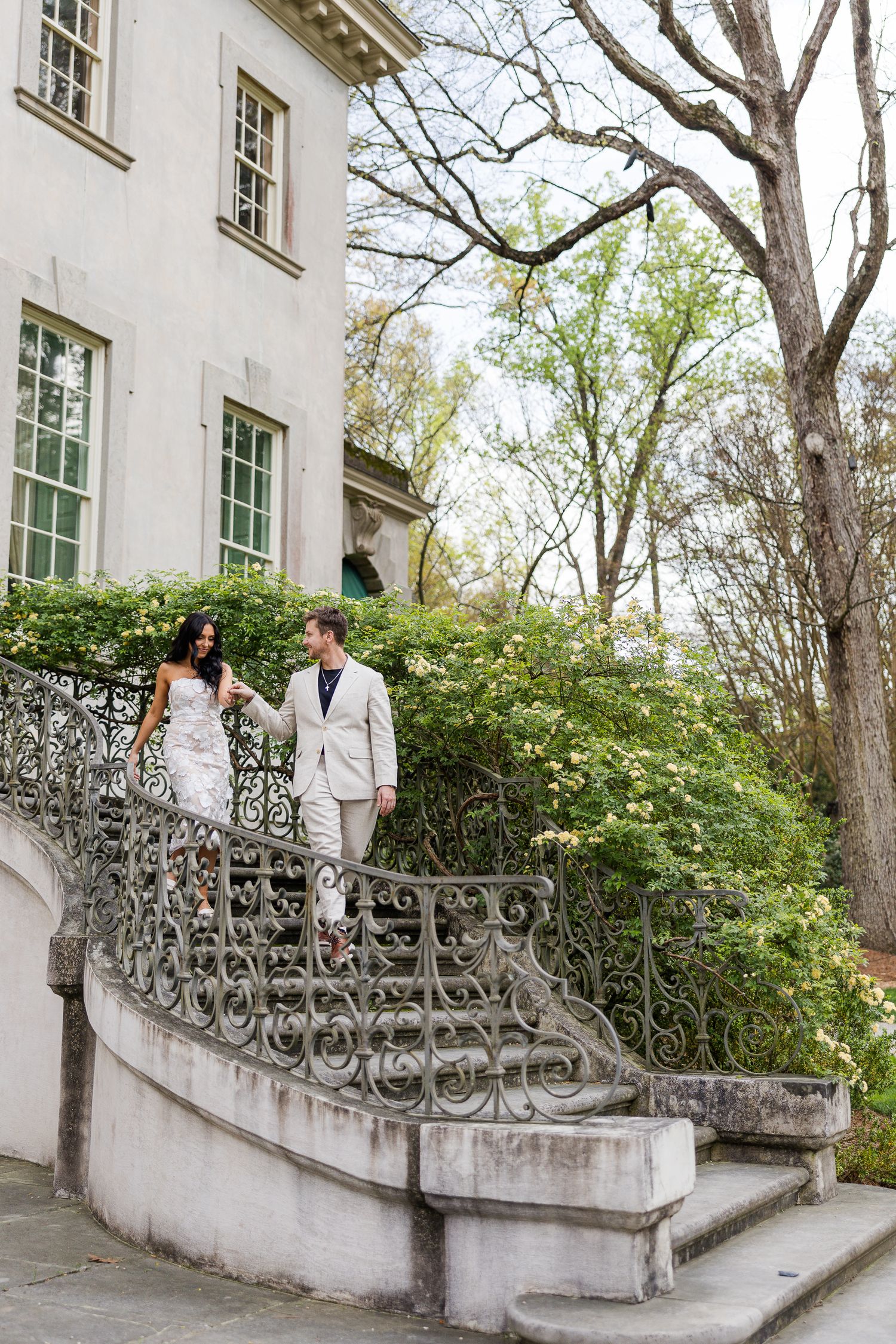 Bride and groom walking down ornate iron staircase at elegant white mansion surrounded by lush greenery.