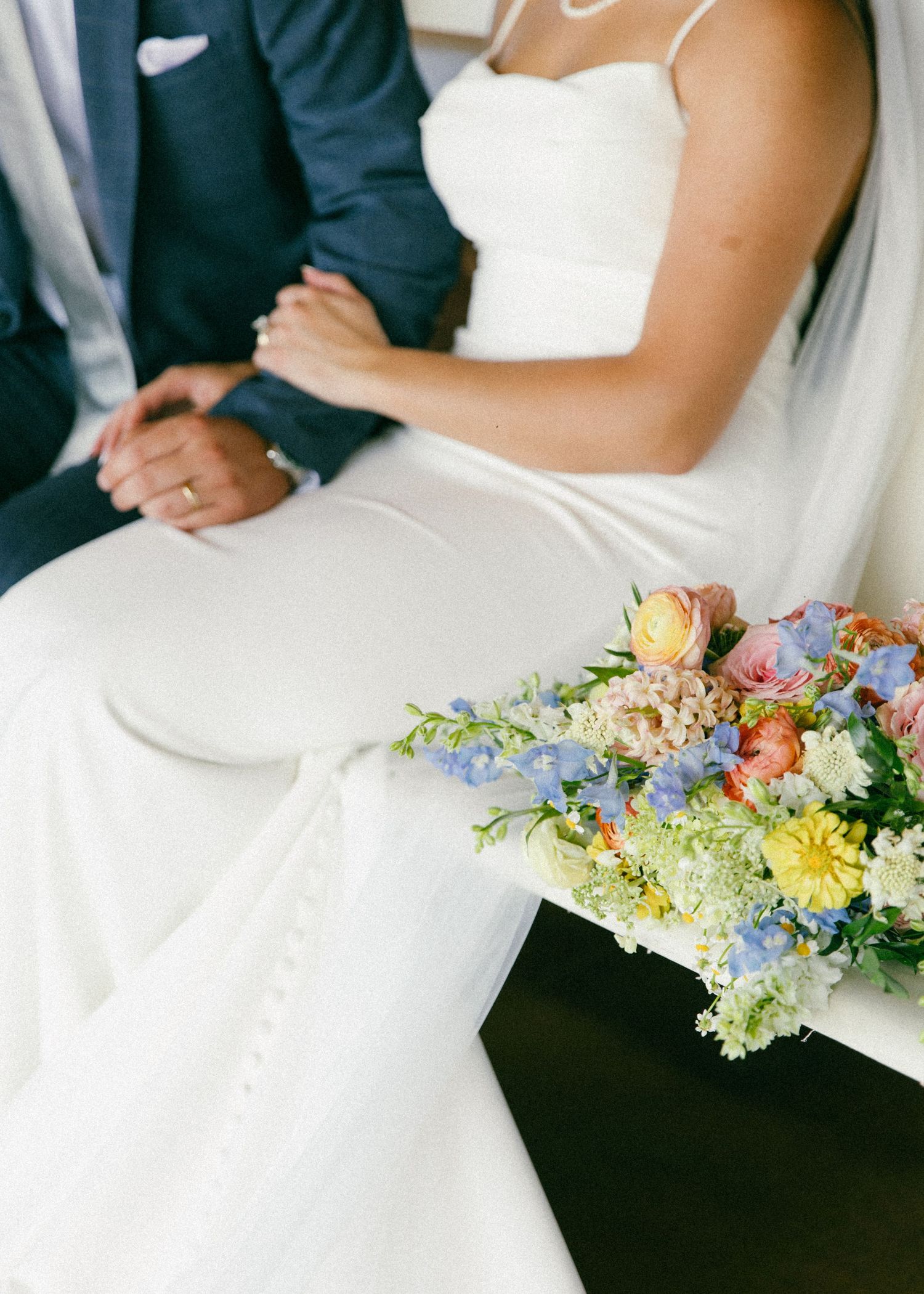 Bride and groom sitting together holding hands, with a colorful spring wedding bouquet of blue, pink, and yellow flowers.