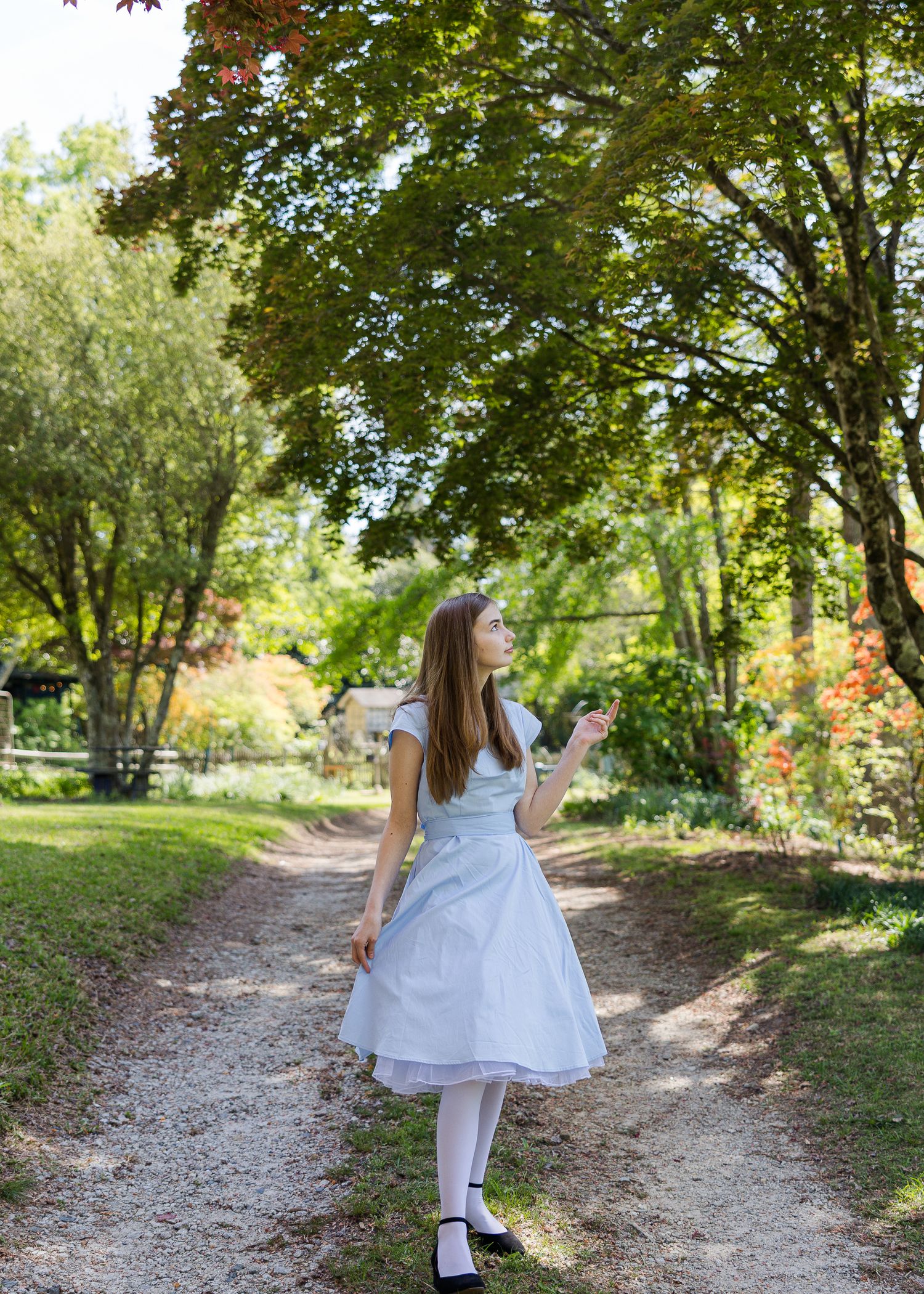 A young girl in a white dress and tights walks along a garden path surrounded by lush green trees.