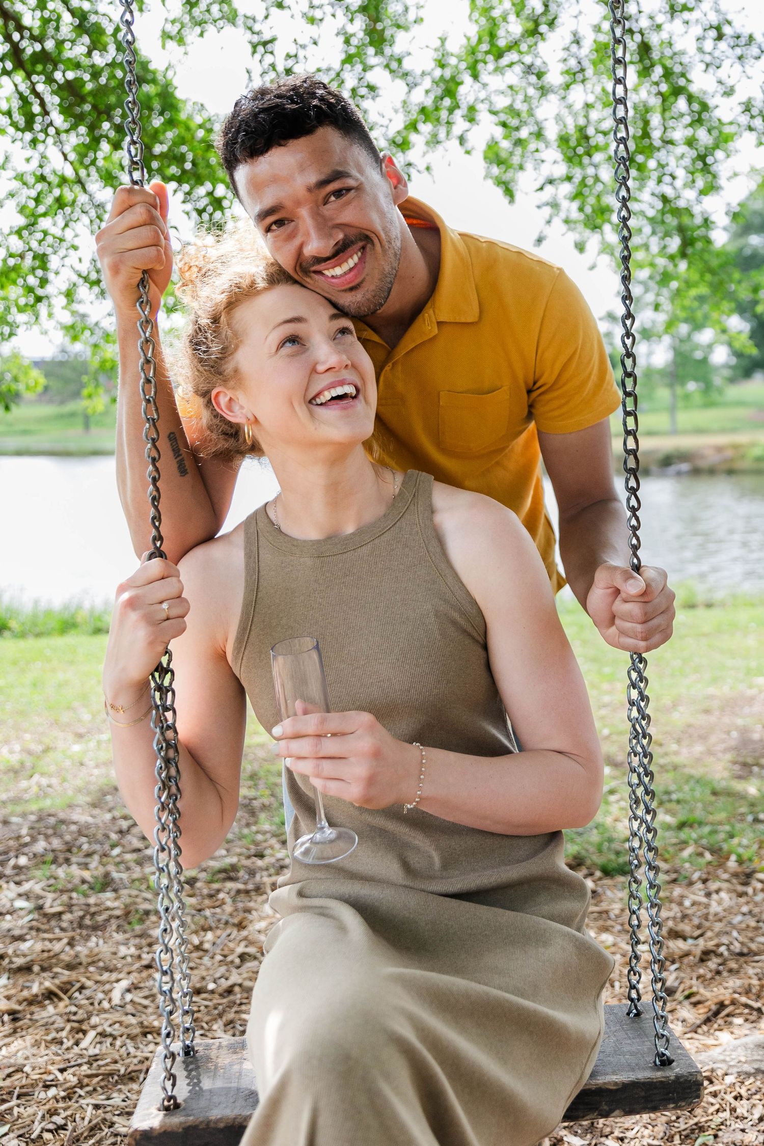 Happy couple enjoying a swing in the park, woman holding champagne glass, man in yellow shirt smiling outdoors.