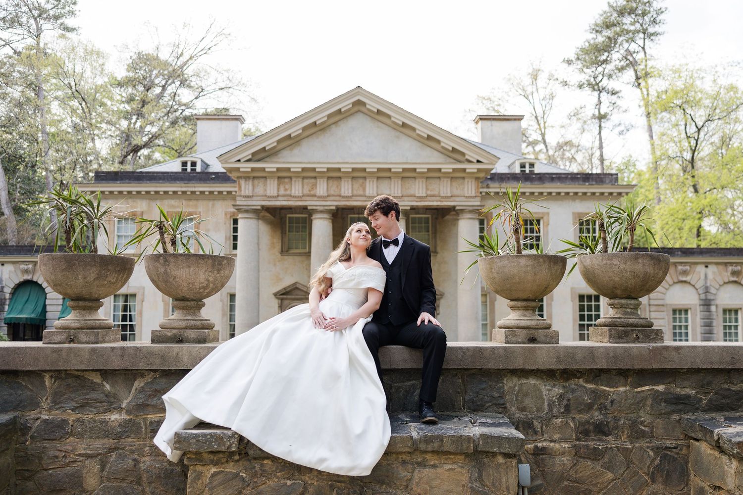 Bride and groom sitting on stone balustrade at elegant Georgian mansion wedding venue surrounded by lush trees.