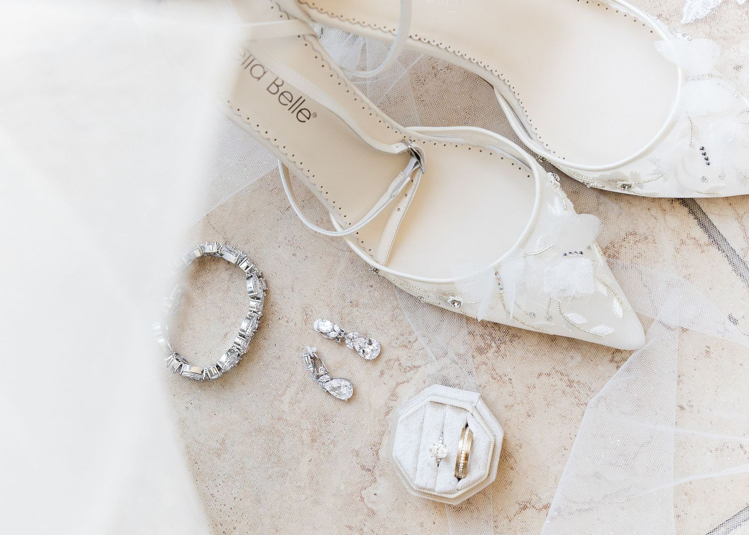 Elegant bridal accessories flat lay with white heels, crystal bracelet, earrings, and gold rings on marble.