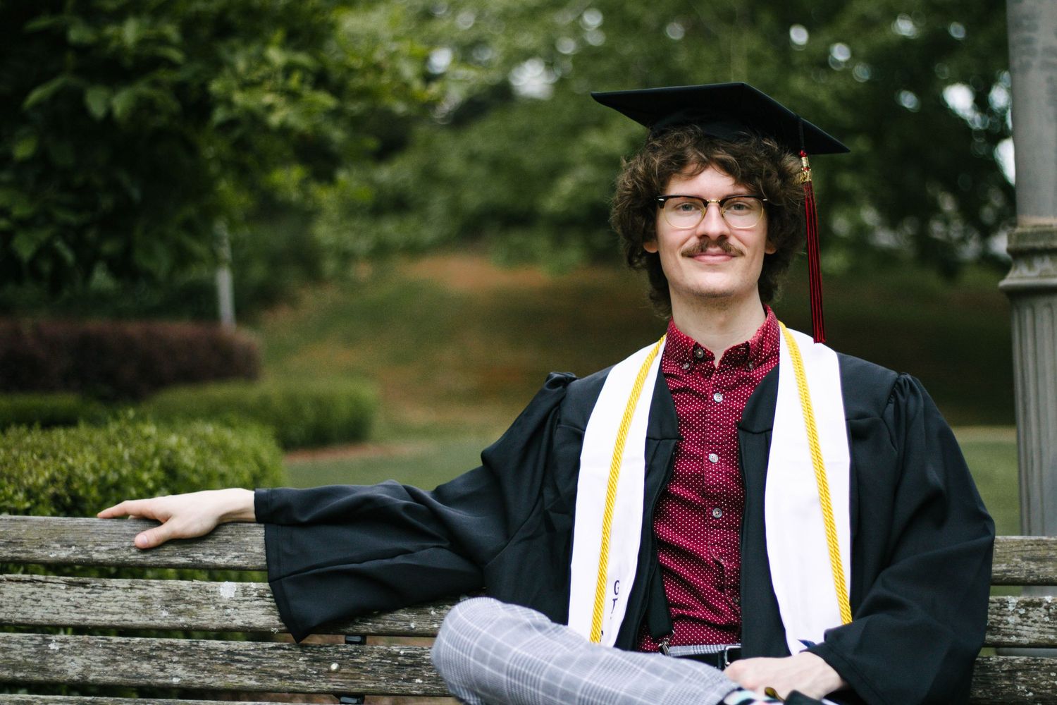 Graduate in burgundy shirt poses with academic stole and honors cords outdoors.