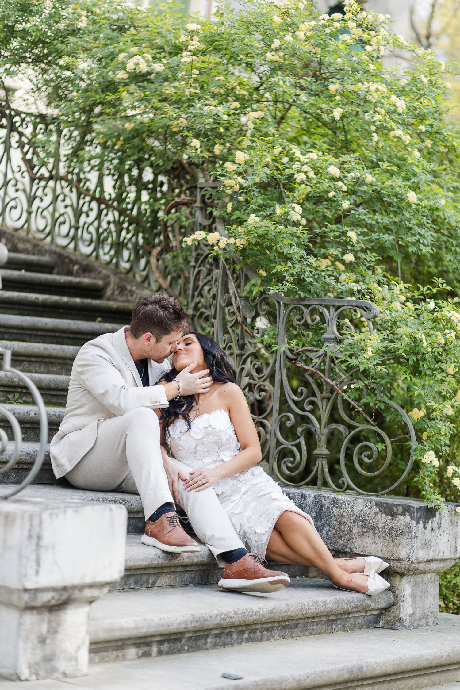 Romantic couple sitting on stone steps near ornate iron railing and blooming bushes, man in suit kissing woman in white dress.