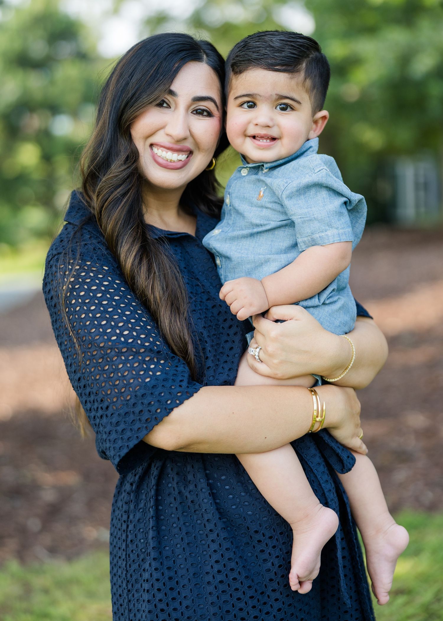 Smiling woman in navy dress holding a happy baby boy outdoors in a park setting.