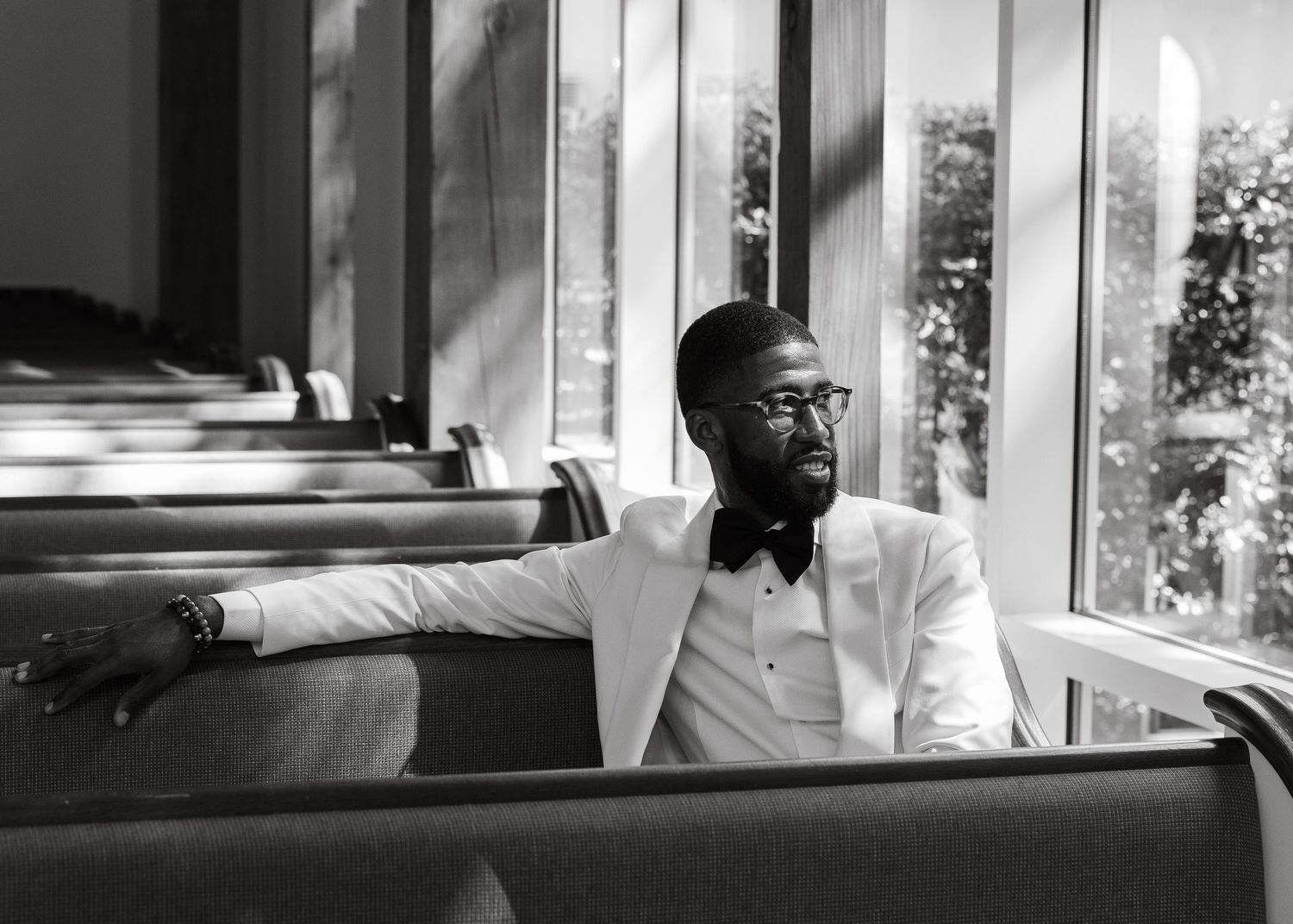 Stylish man in white suit and bow tie sits in church pew, gazing thoughtfully, black and white photo.