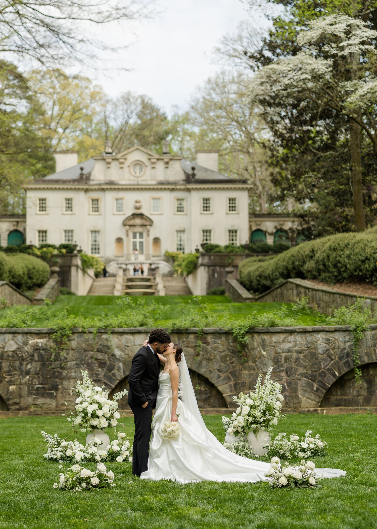 Bride and groom kissing on lush green lawn with white floral arrangements and elegant estate mansion backdrop.