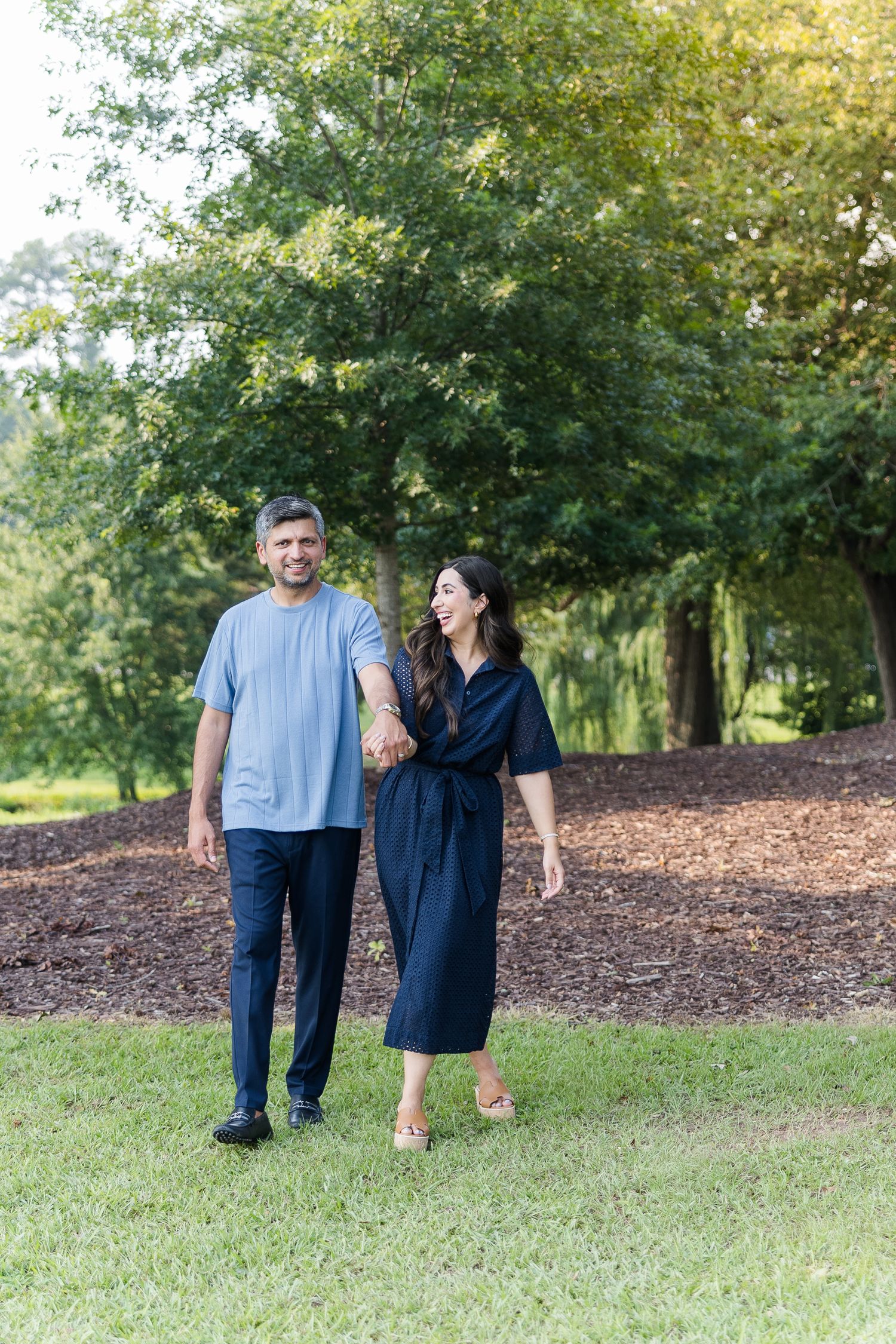 Couple holding hands and walking together in a lush green park, wearing coordinated navy and blue outfits.