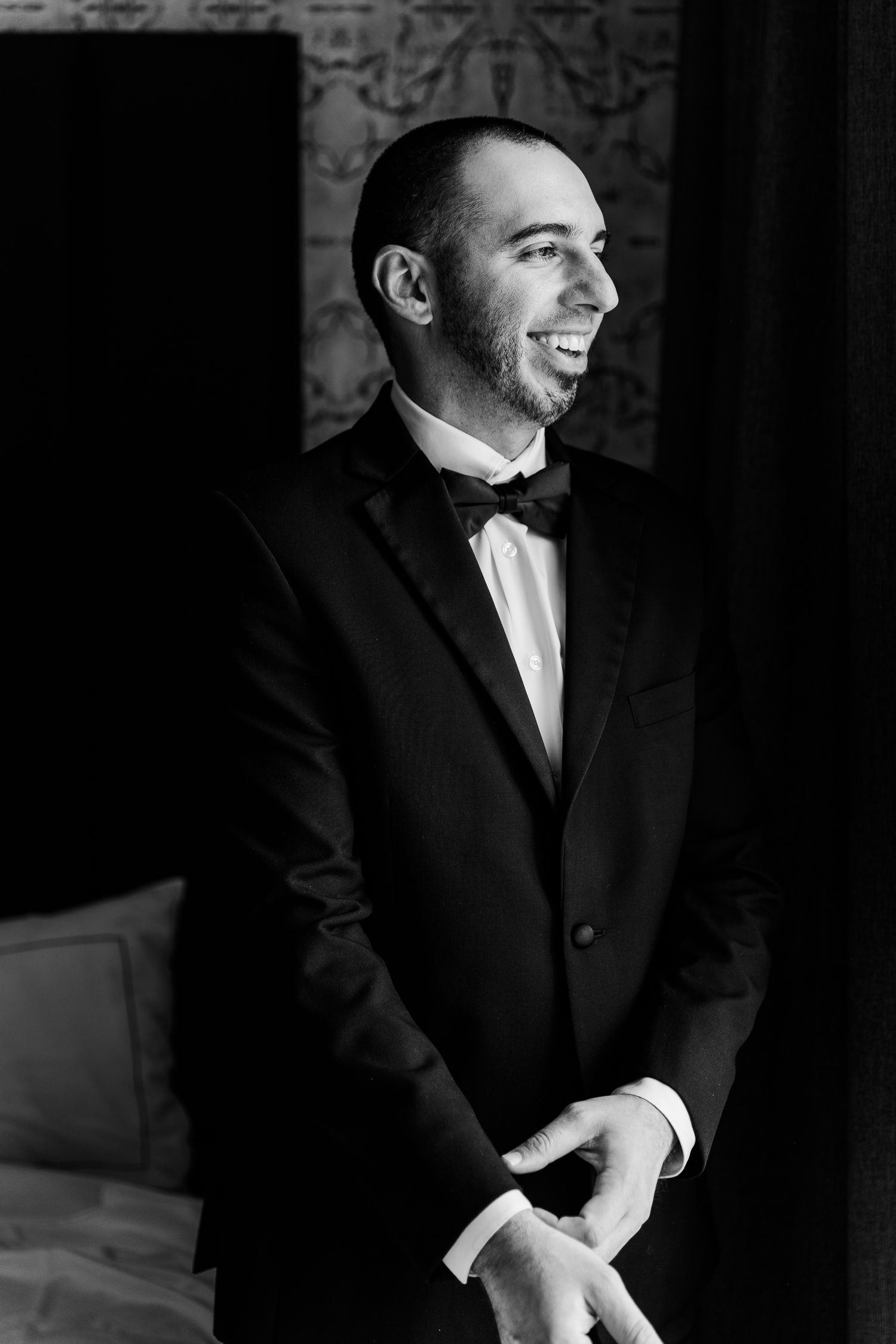 Black and white portrait of a smiling groom in a tuxedo with bow tie adjusting his cufflinks.