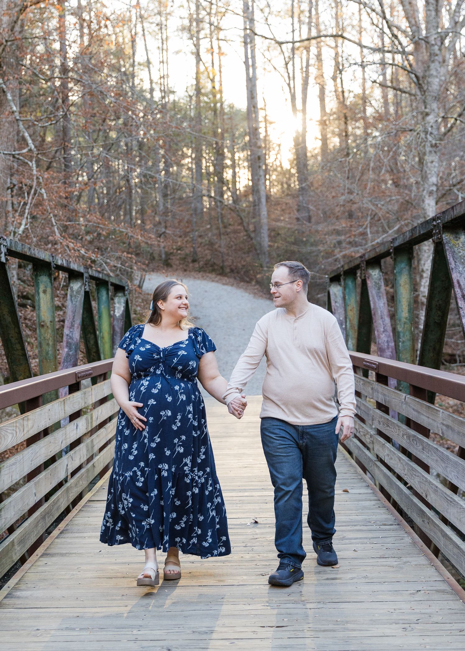 Pregnant woman in navy floral dress holds hands with man on wooden bridge surrounded by autumn trees at sunset.