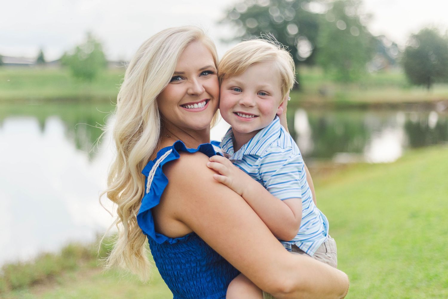 Smiling blonde woman in blue dress holding a young blond boy near a pond on a green lawn.