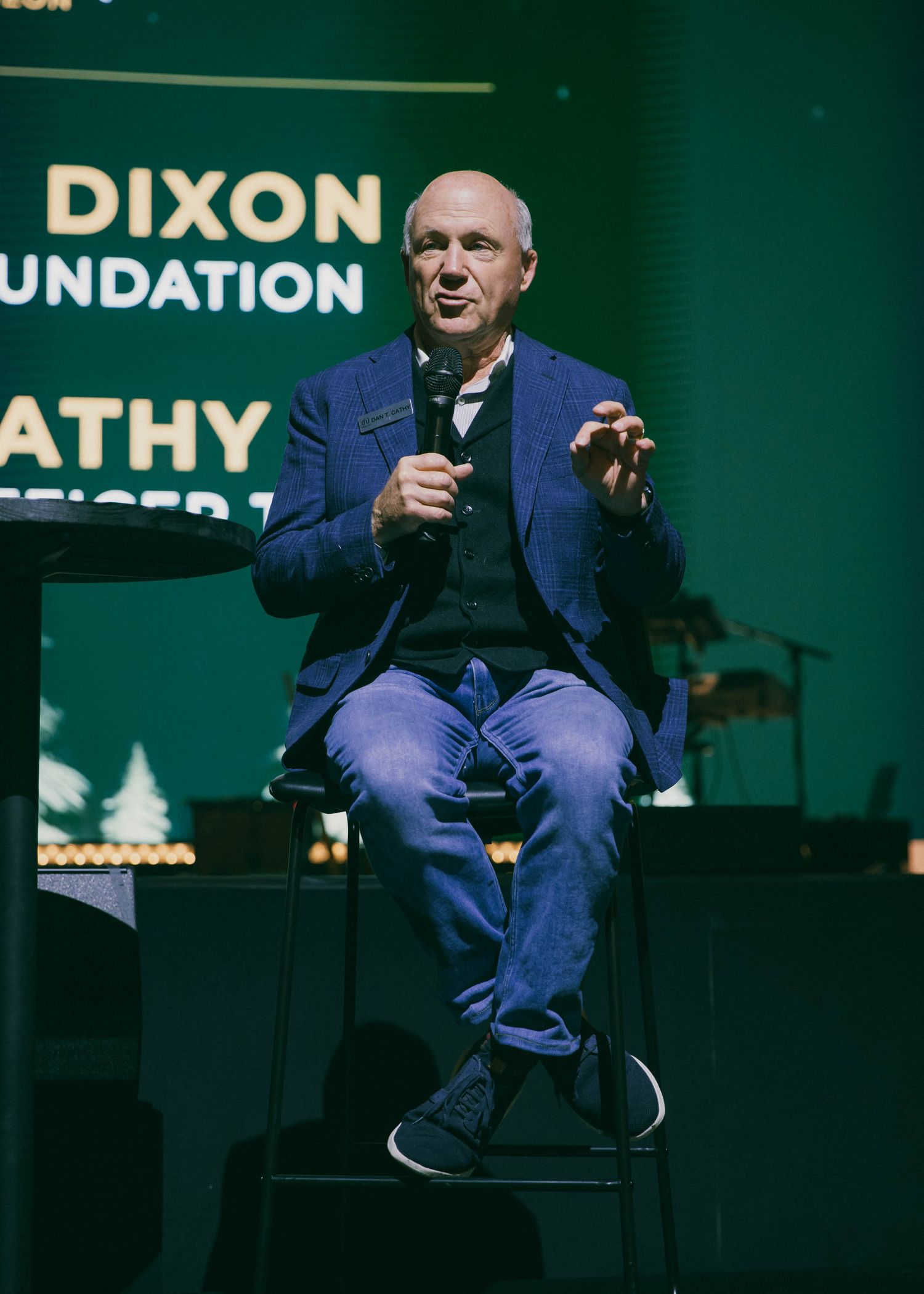 Speaker seated on stage during presentation with branded backdrop.