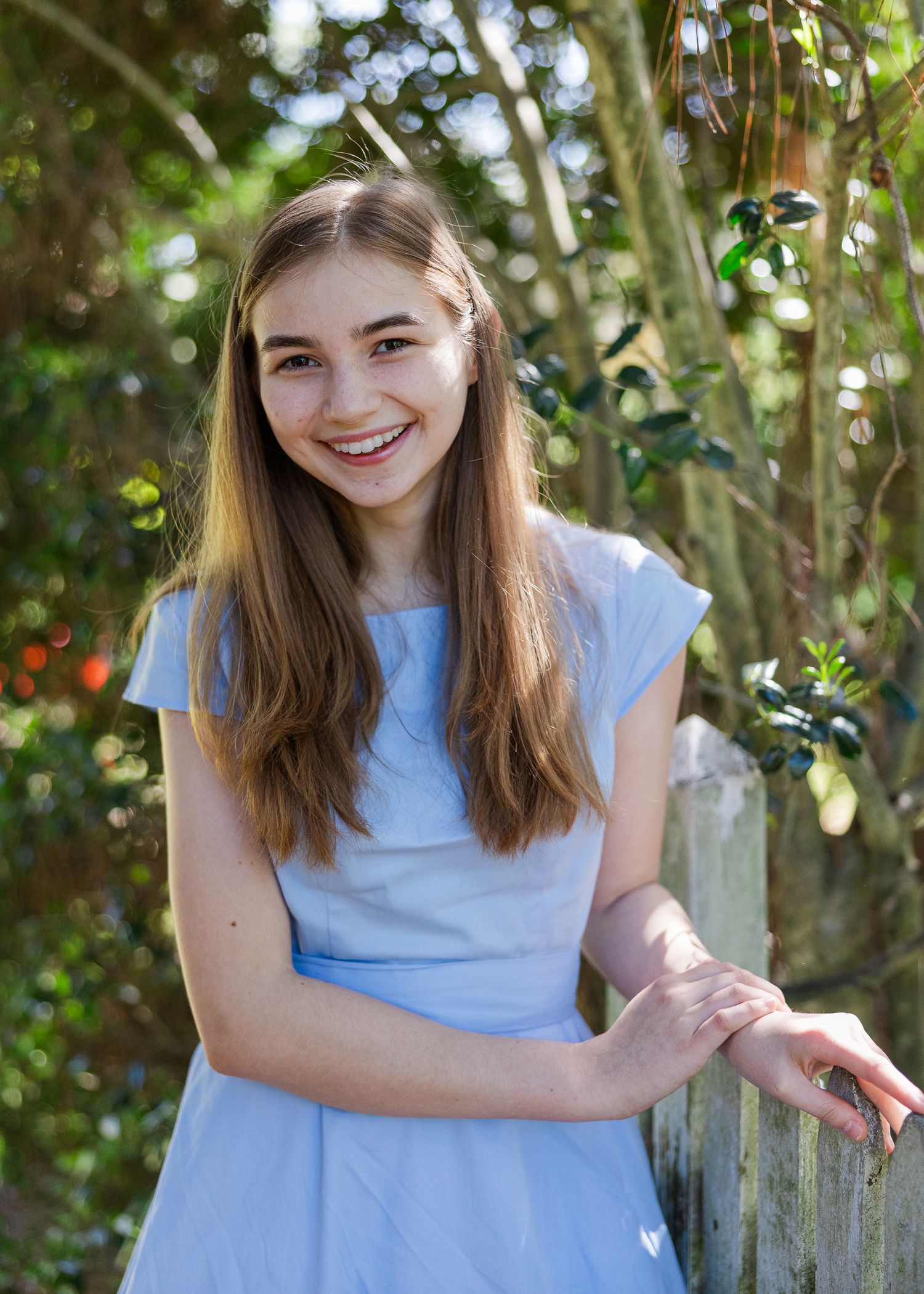 Smiling teenage girl in a light blue dress leaning on a wooden fence outdoors among green trees.