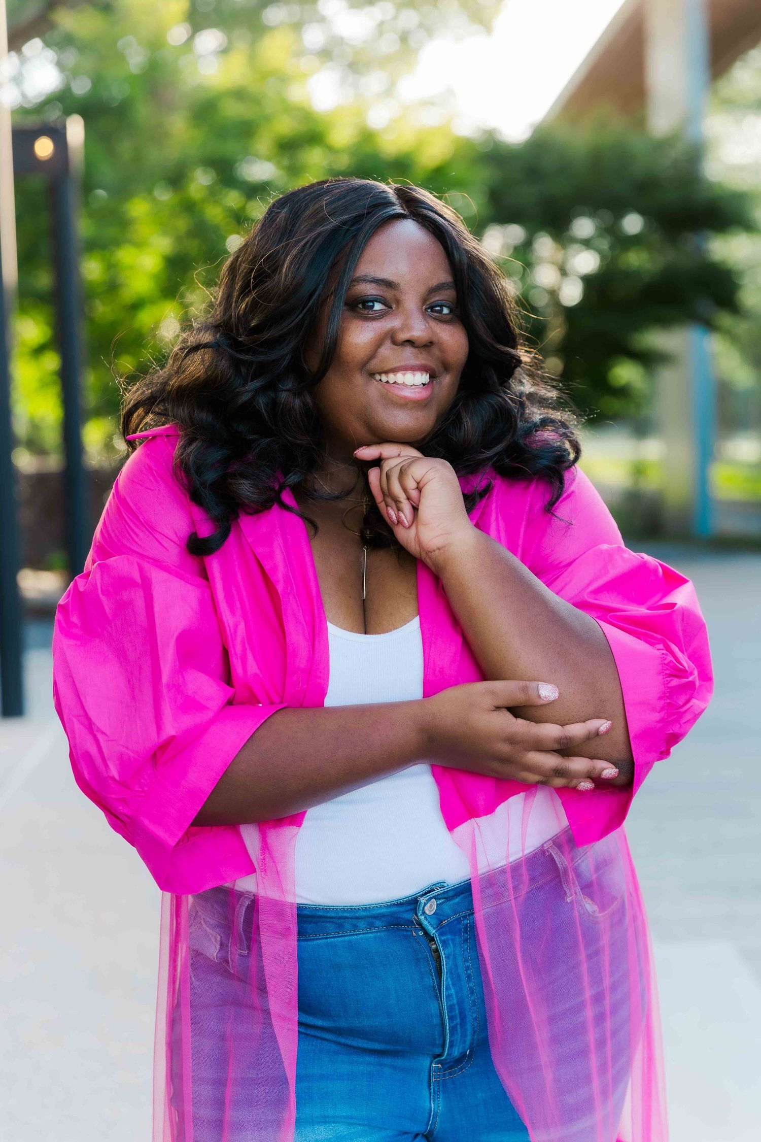 Smiling young Black woman in pink jacket and jeans posing outdoors with hand on chin.