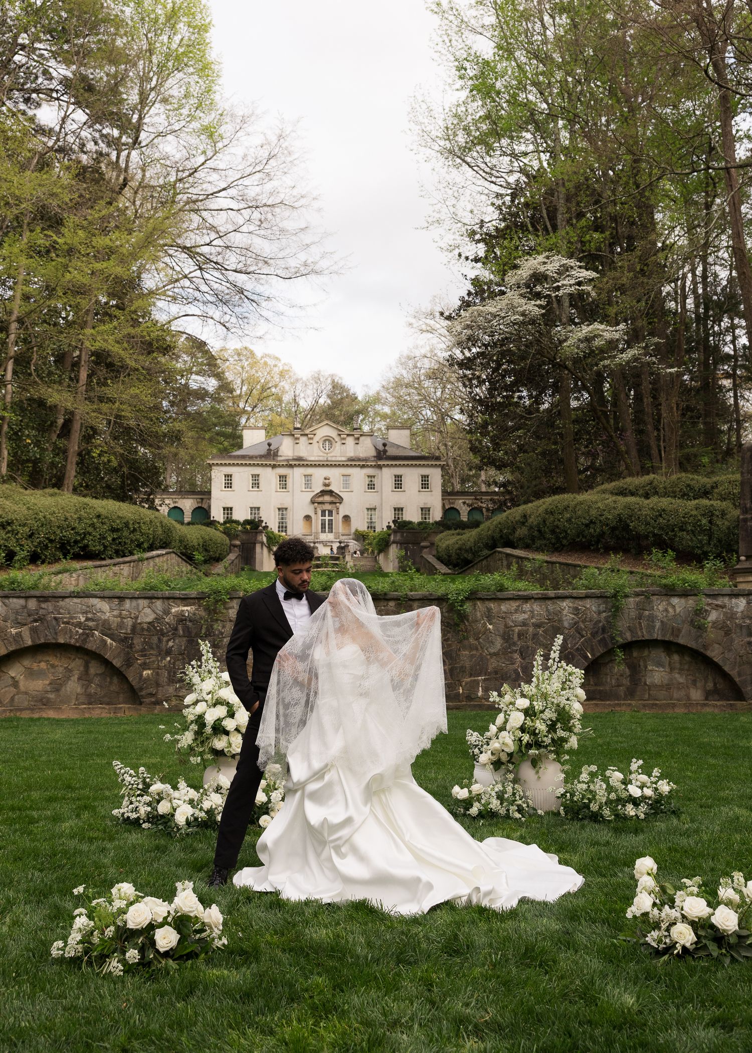 Bride and groom share a romantic moment on lush lawn with white floral arrangements and elegant manor backdrop.