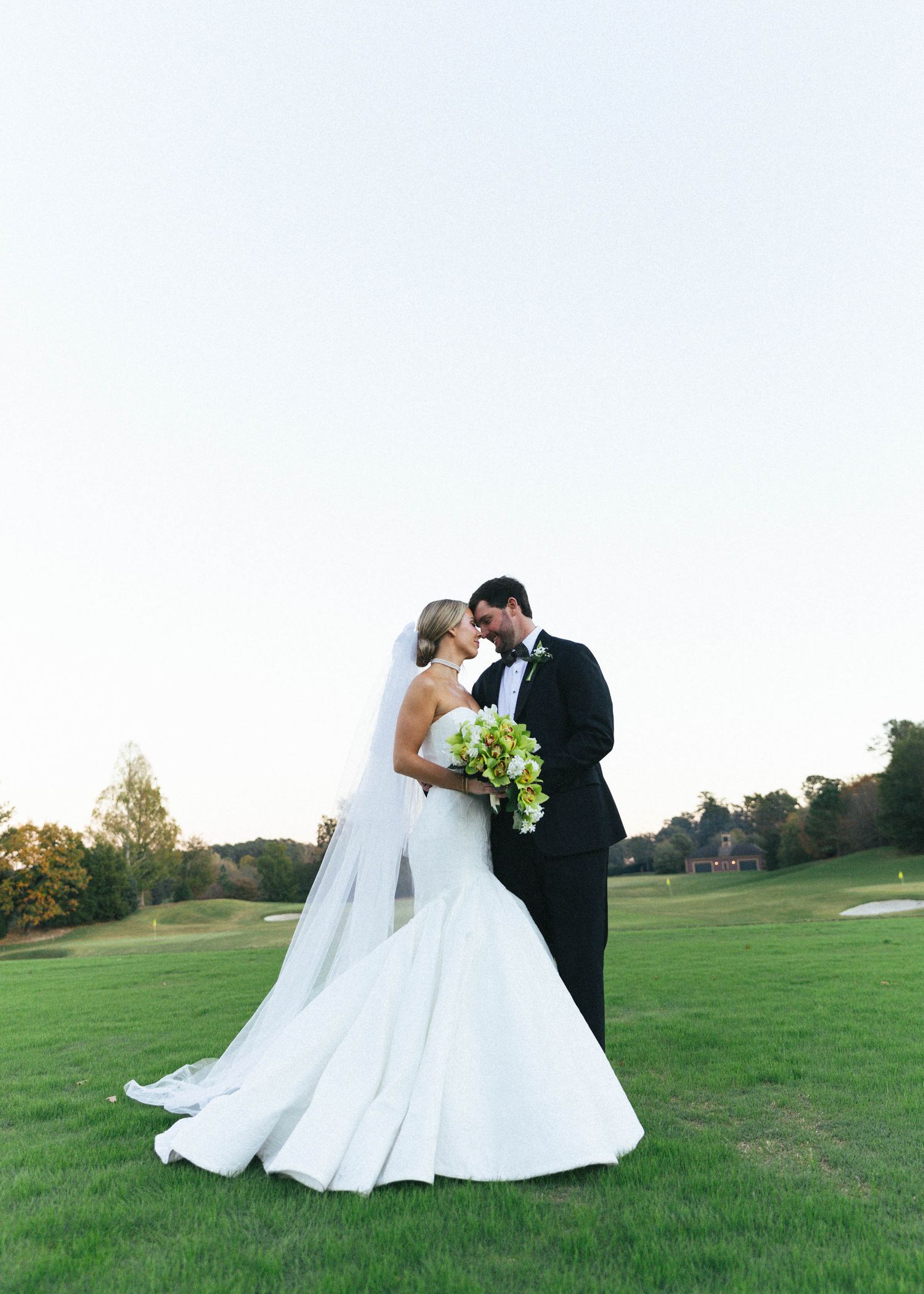 Bride and groom share a romantic moment on a golf course at dusk, bride in mermaid gown with veil holding green bouquet.