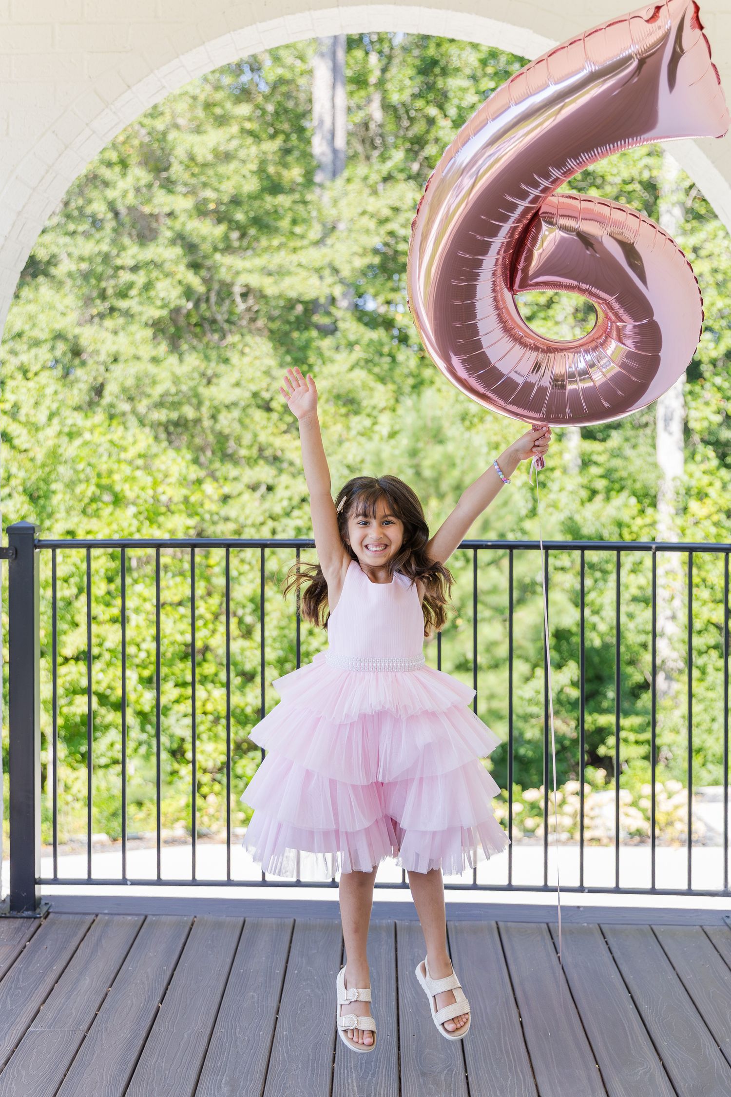 A joyful girl in a pink tulle dress jumps with arms raised, holding a rose gold number 6 balloon outdoors.