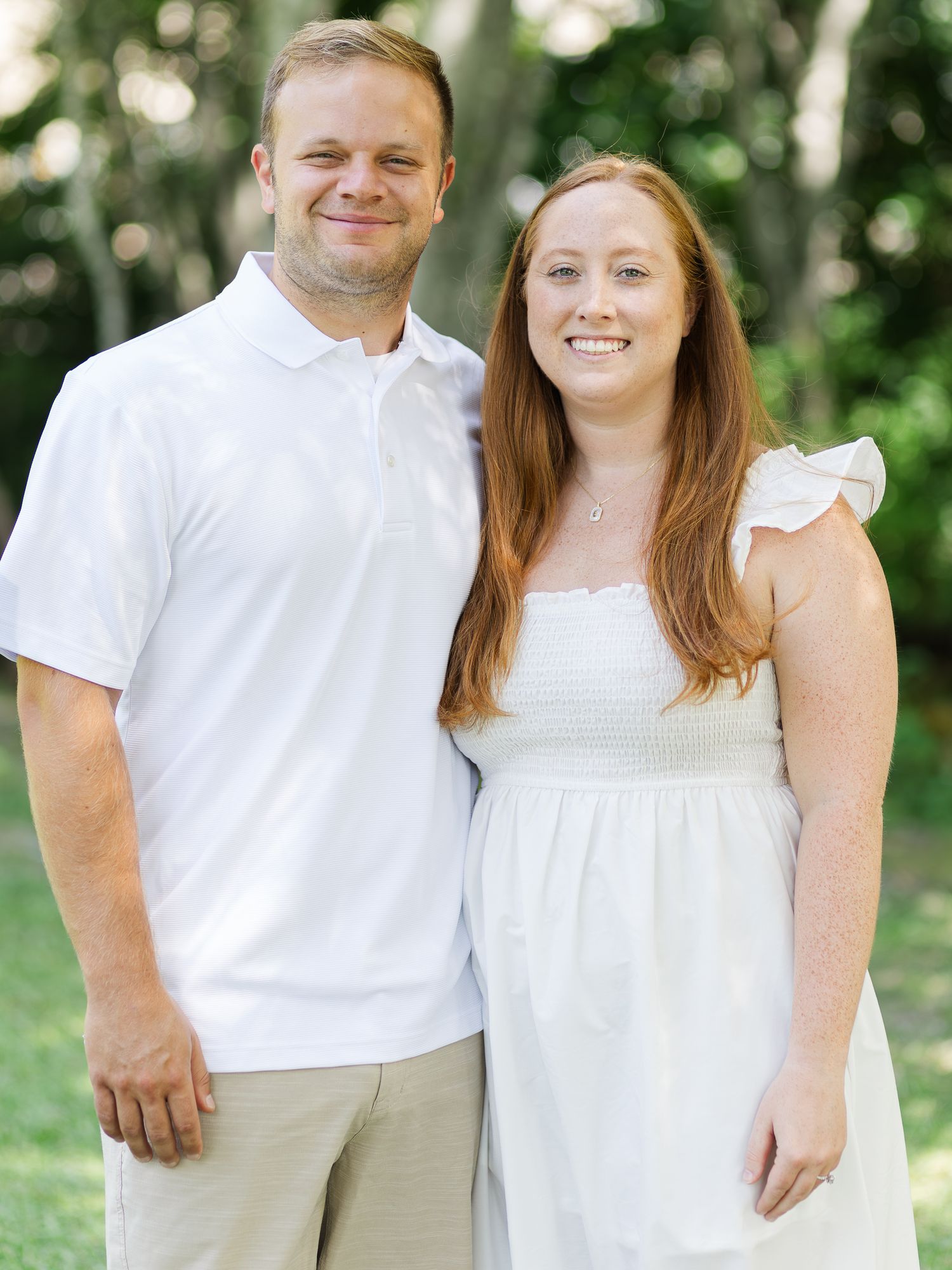 A smiling couple in matching white outfits poses together outdoors among lush green trees.