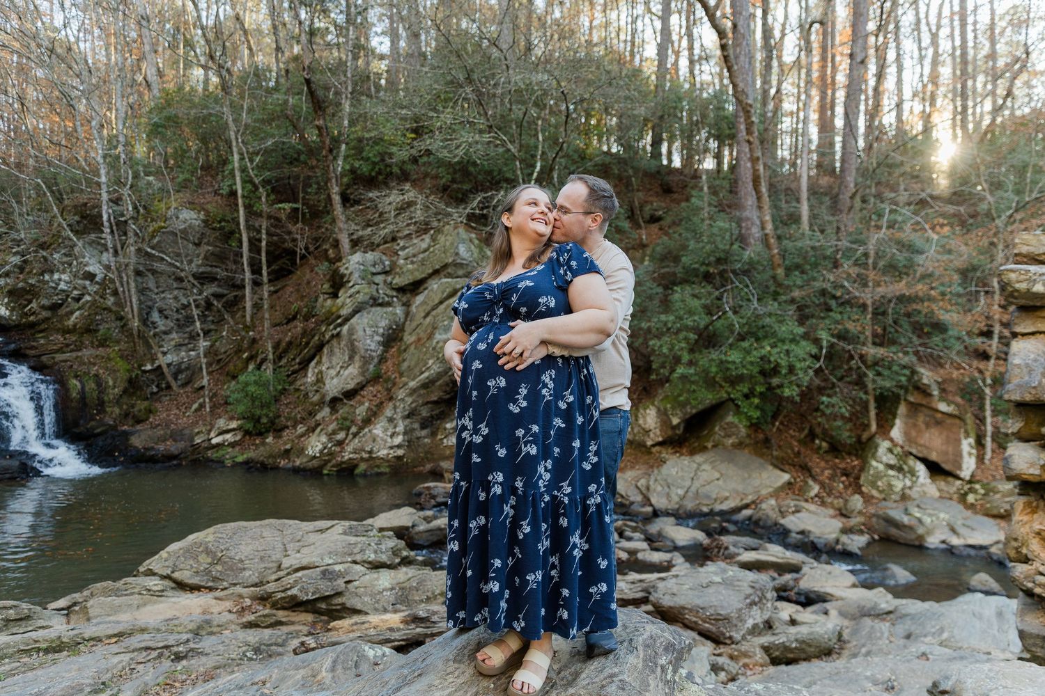 Couple embracing on rocks near a waterfall surrounded by autumn trees in a scenic outdoor engagement photo.