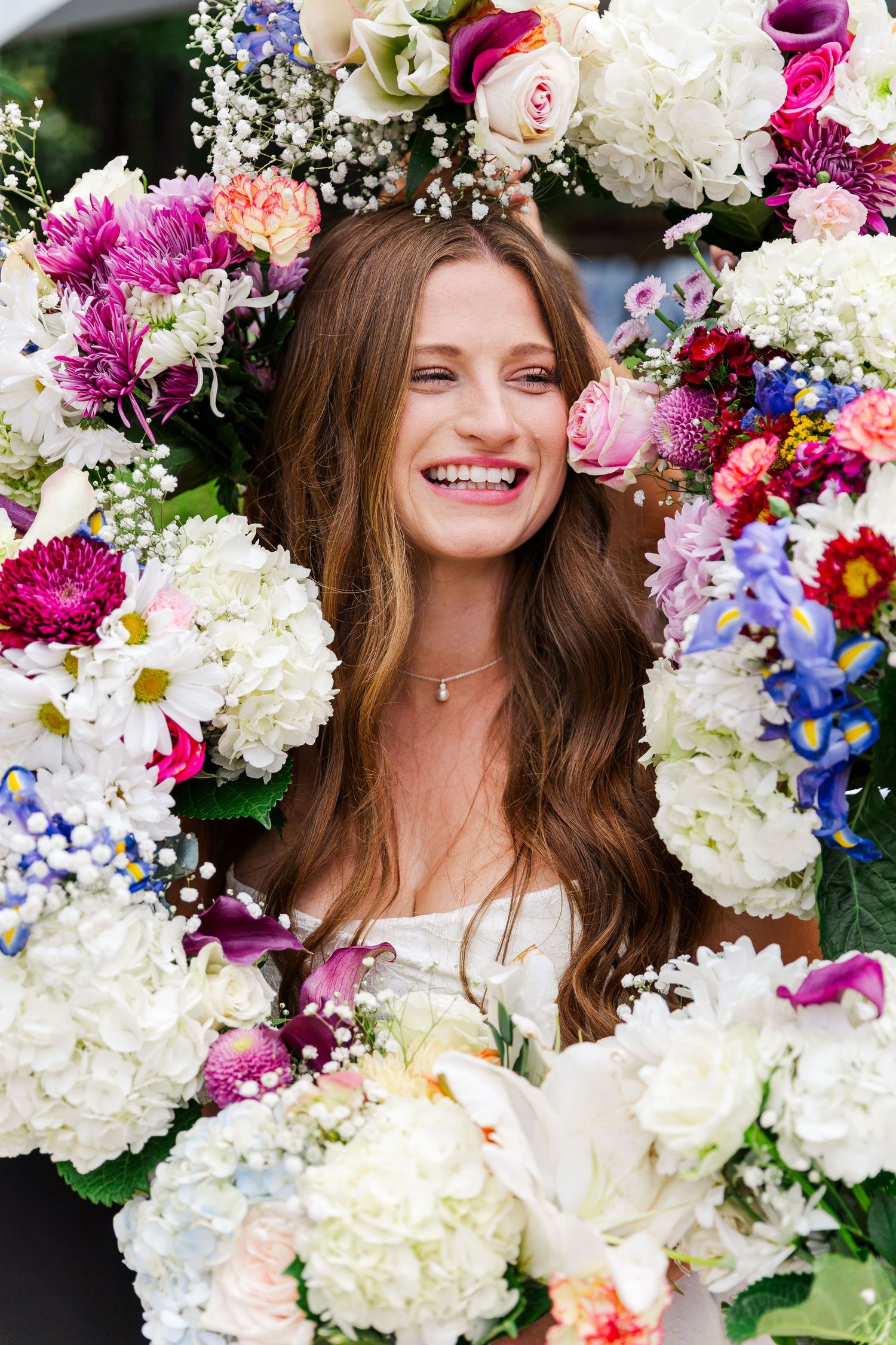 Smiling young woman with long hair framed by a large colorful floral wreath with white, pink, and blue flowers.