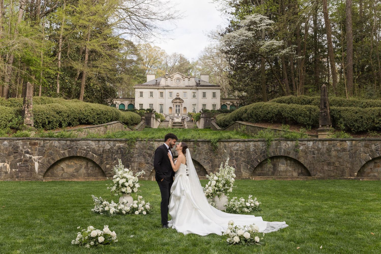 Bride and groom share a romantic moment on lush green lawn before an elegant stone mansion with manicured hedges.