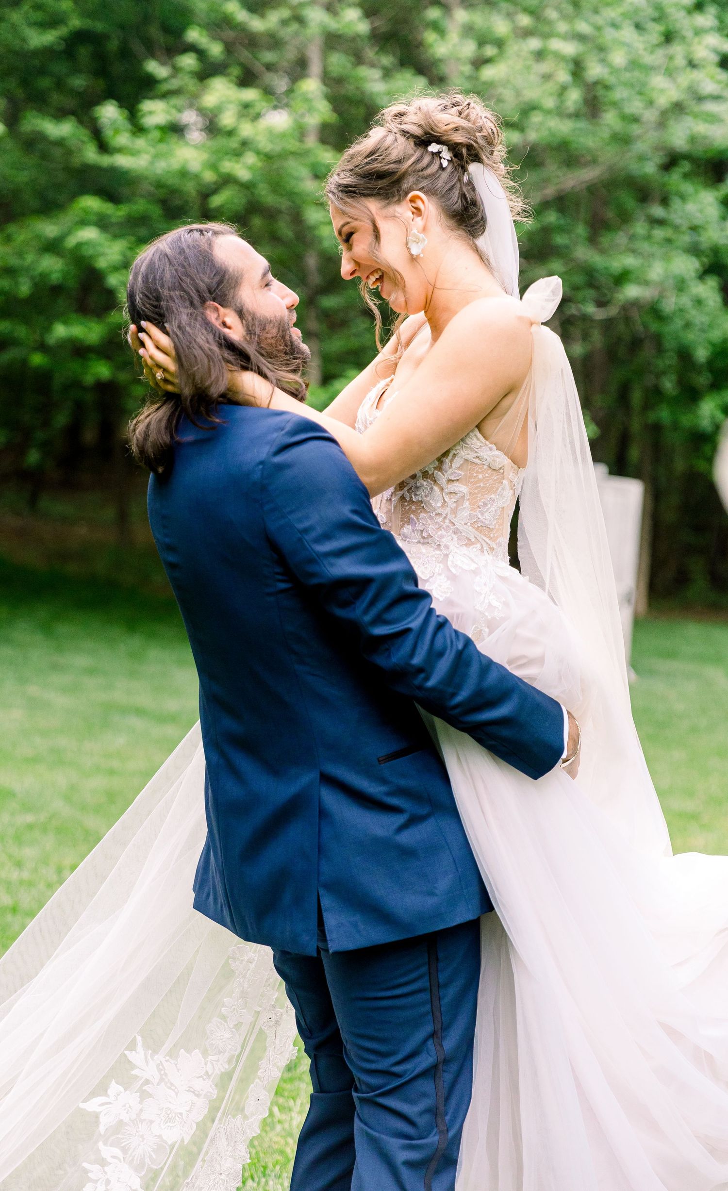 A joyful bride in a lace gown is lifted by her groom in a navy suit, laughing together outdoors on their wedding day.