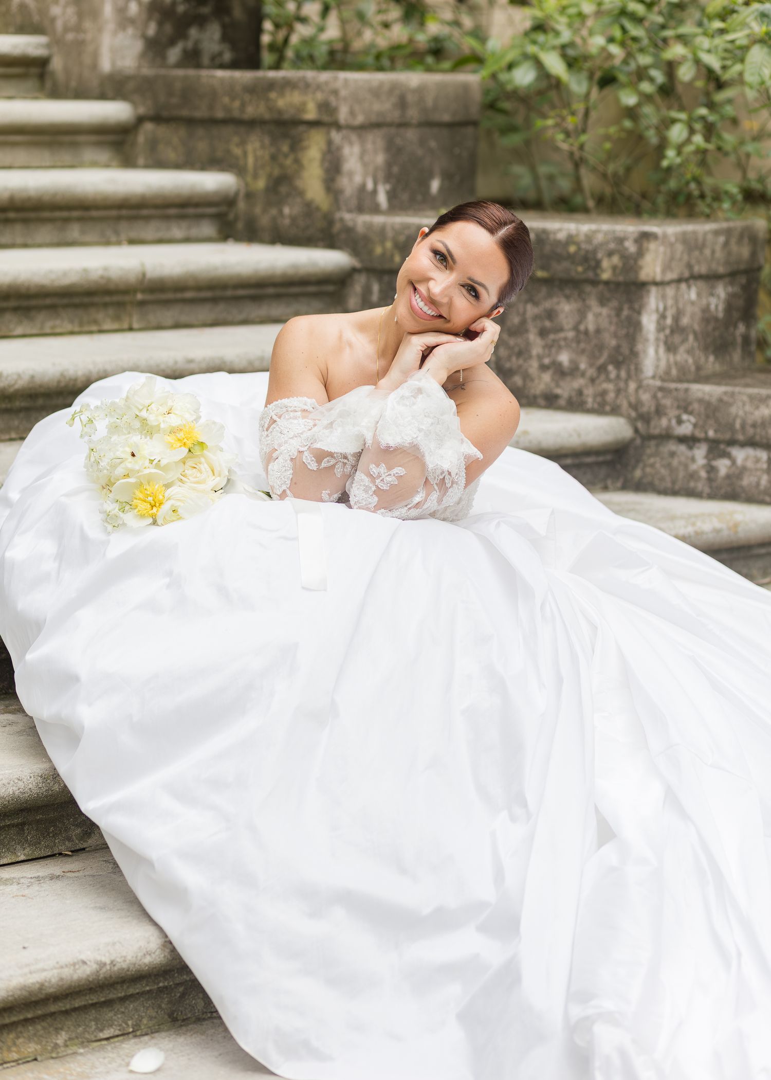 Bride in a white ball gown sits on stone steps, smiling, holding a yellow and white floral bouquet.