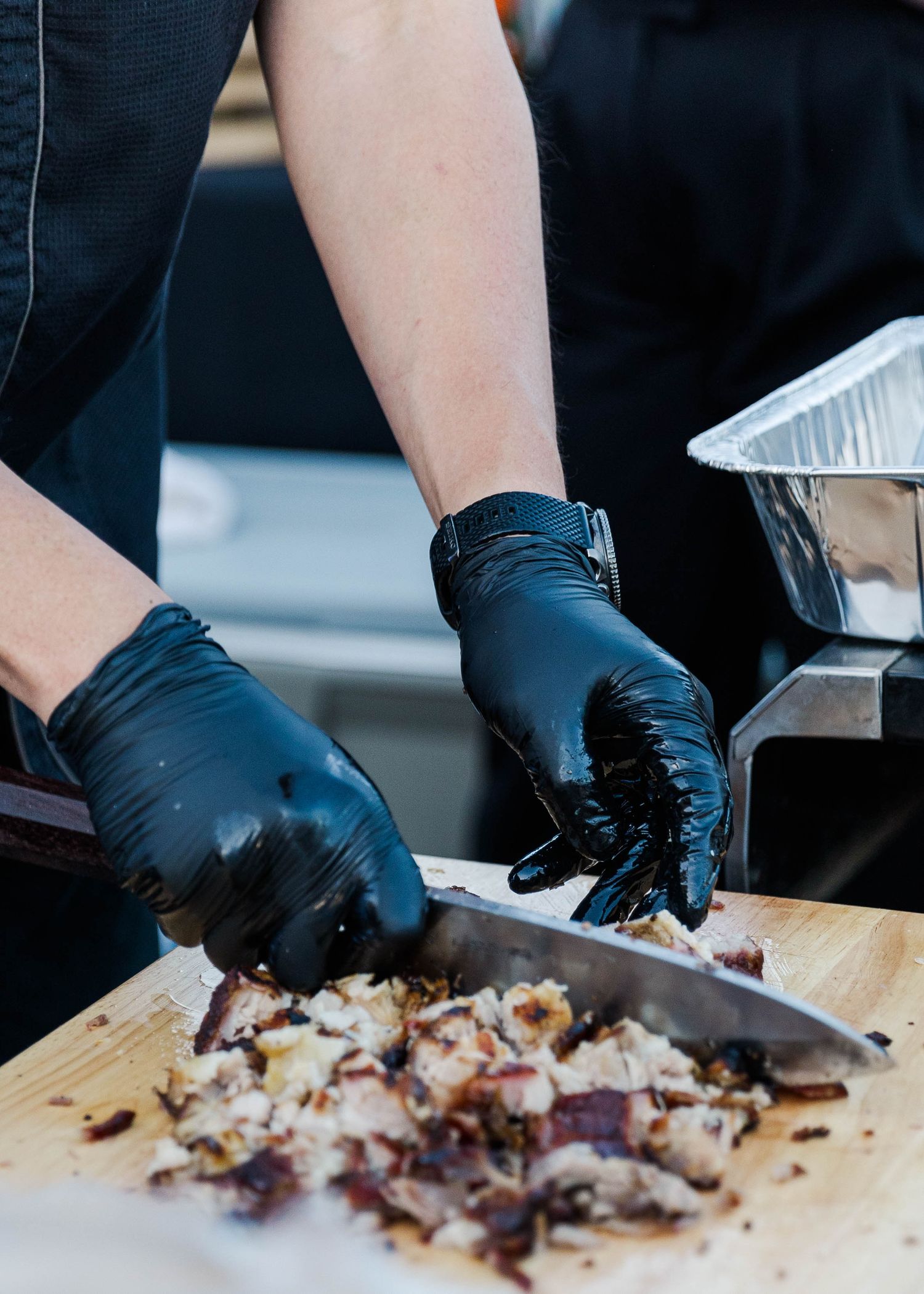 Hands wearing black gloves chopping ingredients on a wooden cutting board.