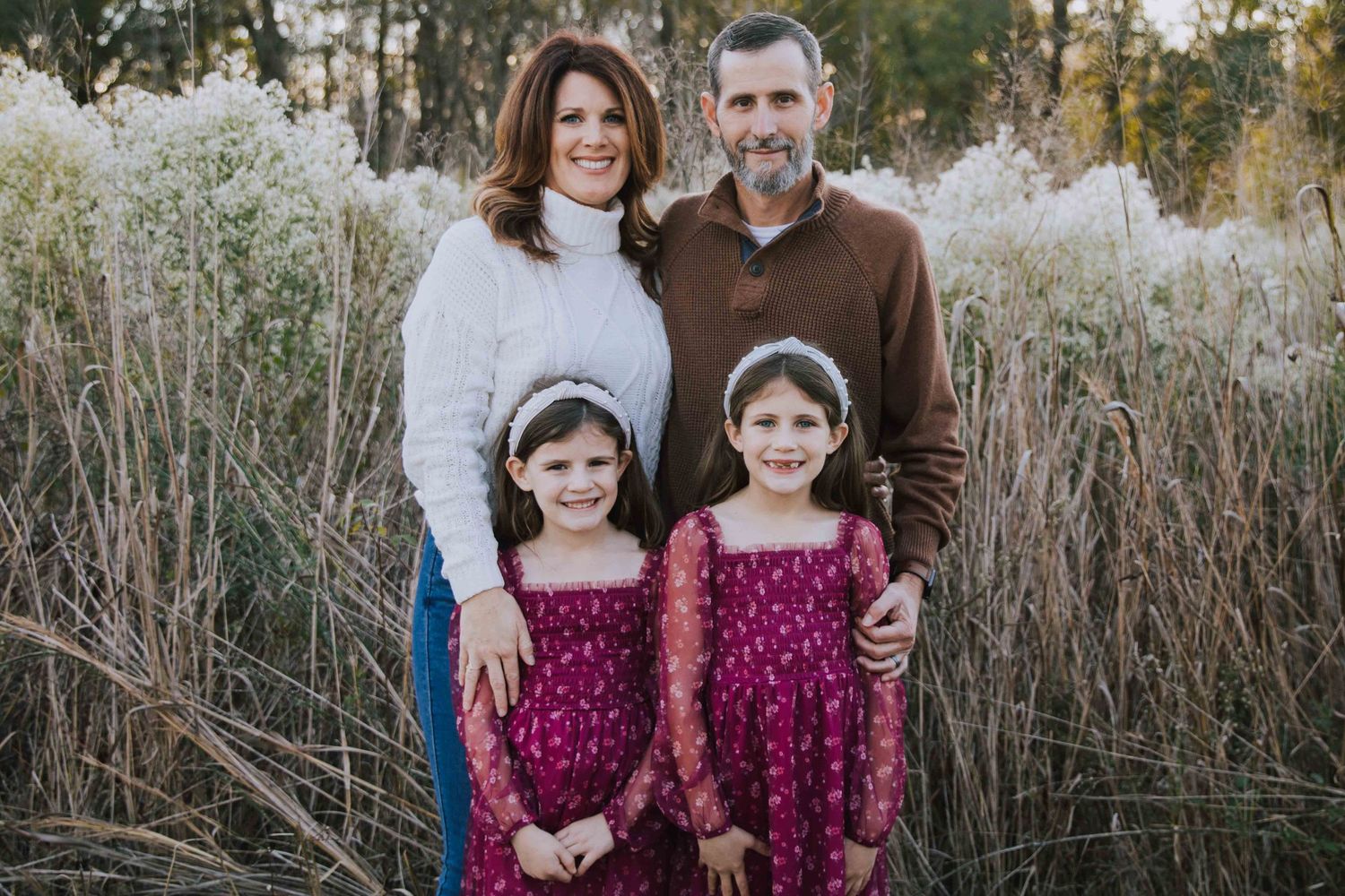 Family of four posing outdoors in fall, parents with two young girls in matching burgundy dresses among tall grasses.