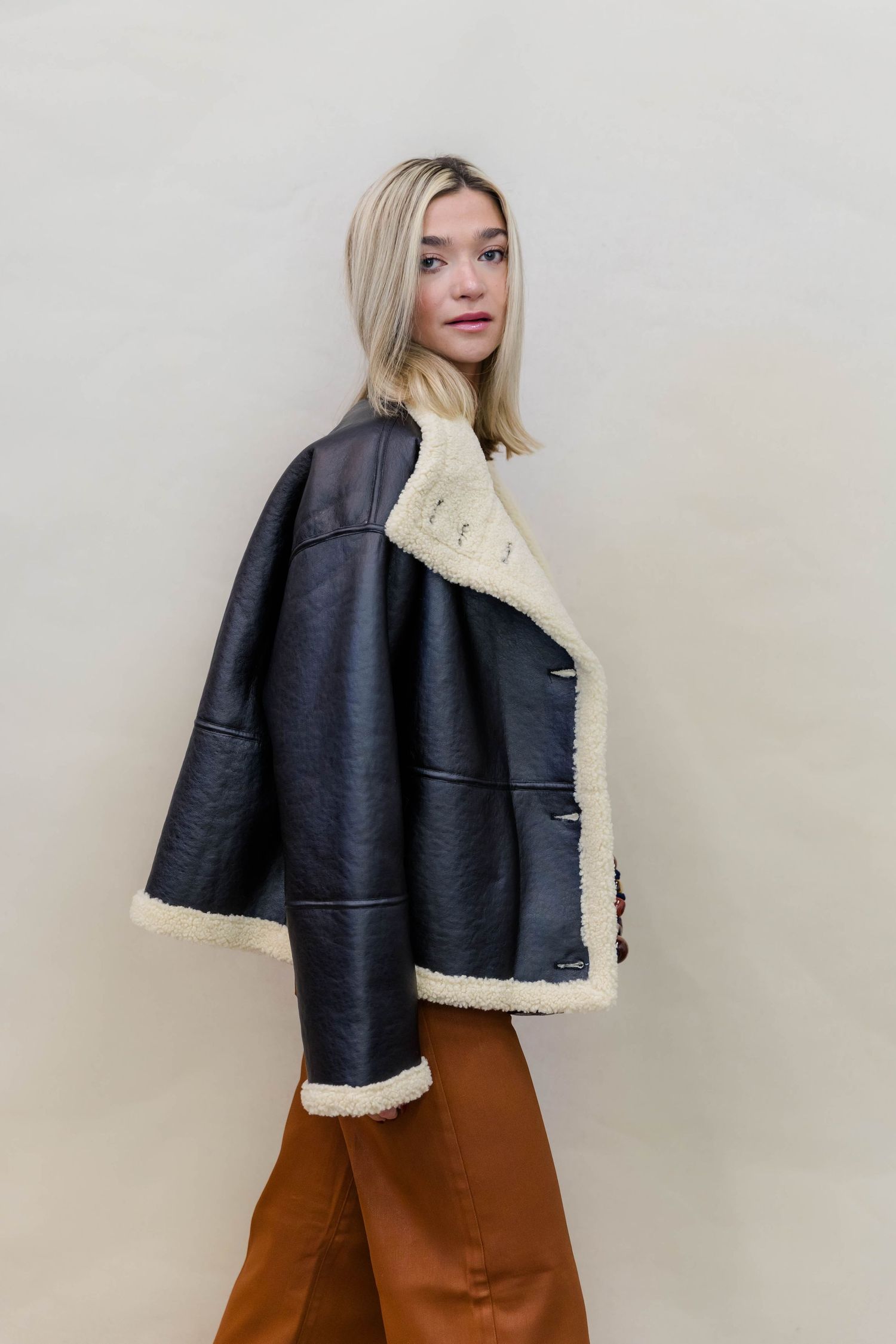 A black leather jacket with cream sherpa lining styled with brown pants against a light background.