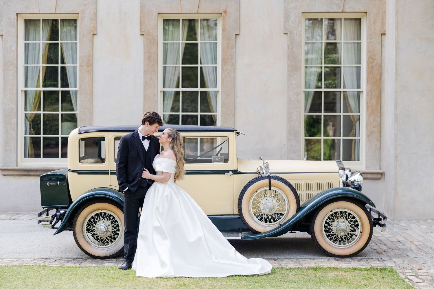 Bride and groom posing together beside a vintage cream and black classic car outside an elegant stone building.