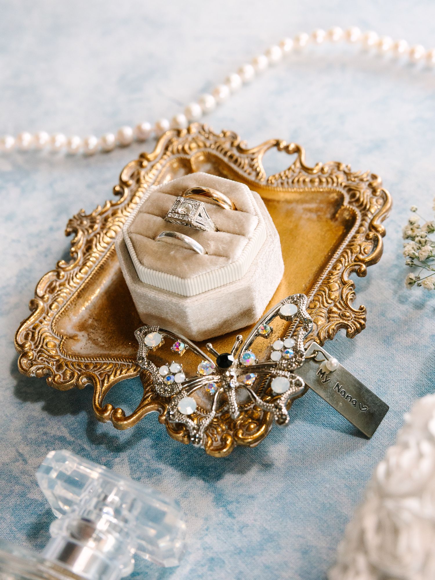 Gold ornate tray with velvet ring box holding wedding rings, pearl necklace, crystal brooch, and perfume bottle.
