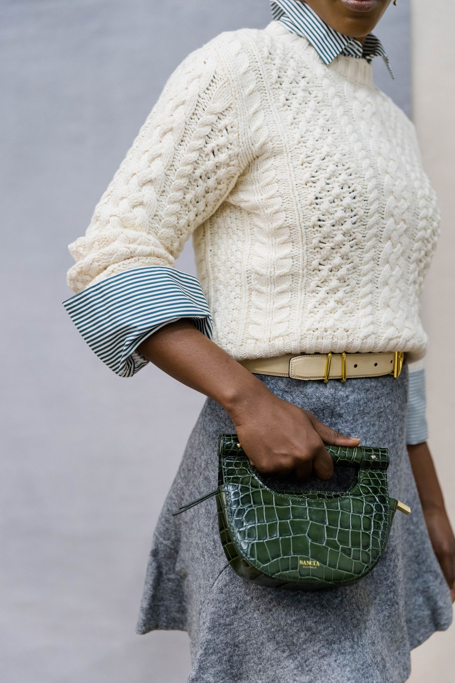 Cream cable knit sweater paired with striped shirt, gray skirt and green crocodile handbag for preppy style.