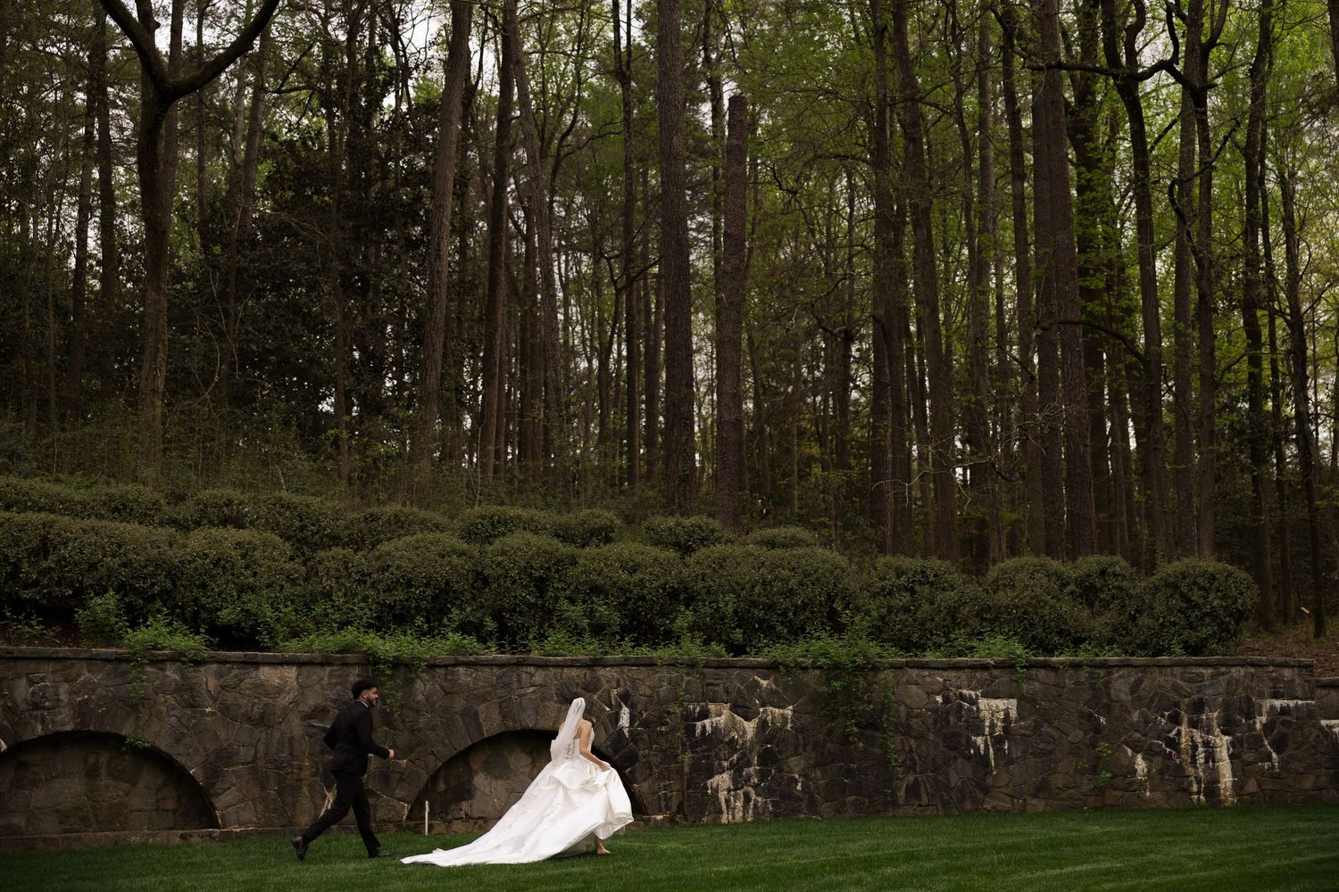 Bride and groom walking on lush green lawn beside stone wall with tall trees in background at outdoor wedding.