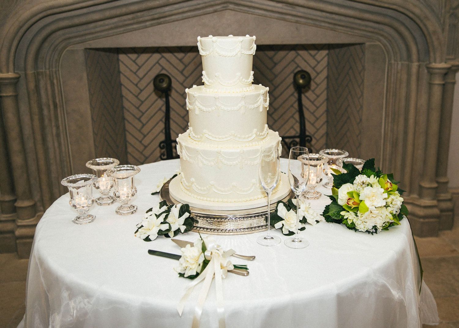 Elegant four-tier white wedding cake on a draped table with floral arrangements and crystal glasses near a fireplace.