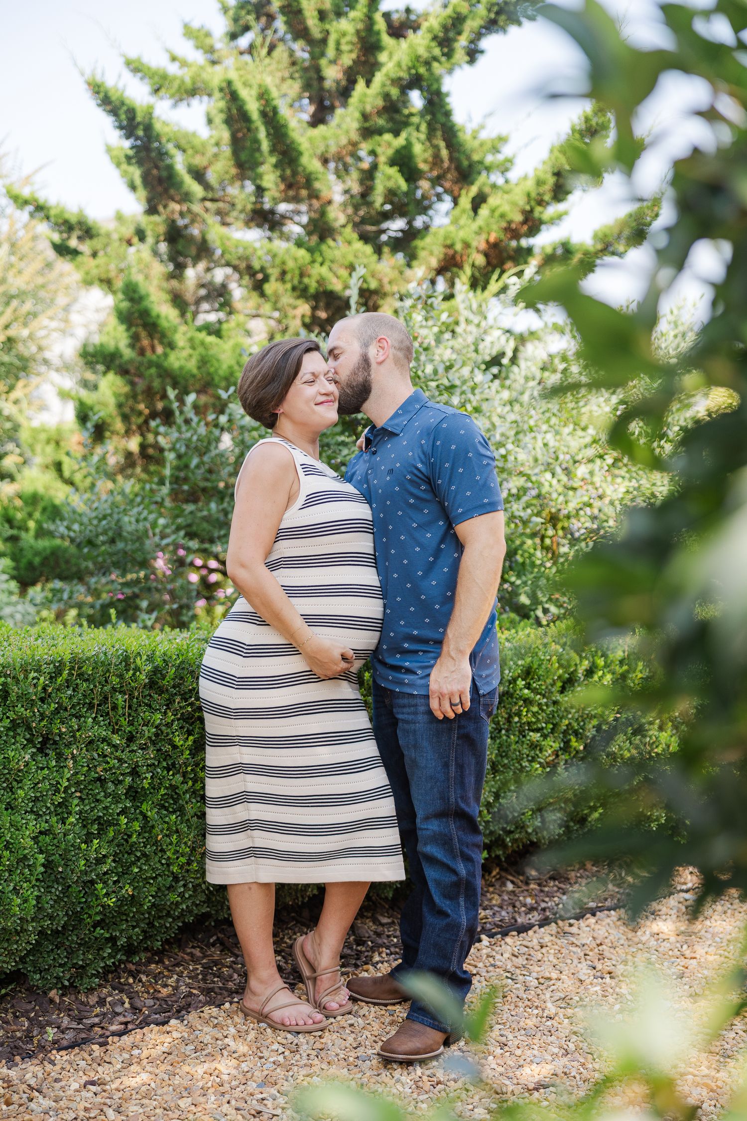 Pregnant woman in striped dress with partner kissing her cheek in lush garden setting during maternity photo shoot.