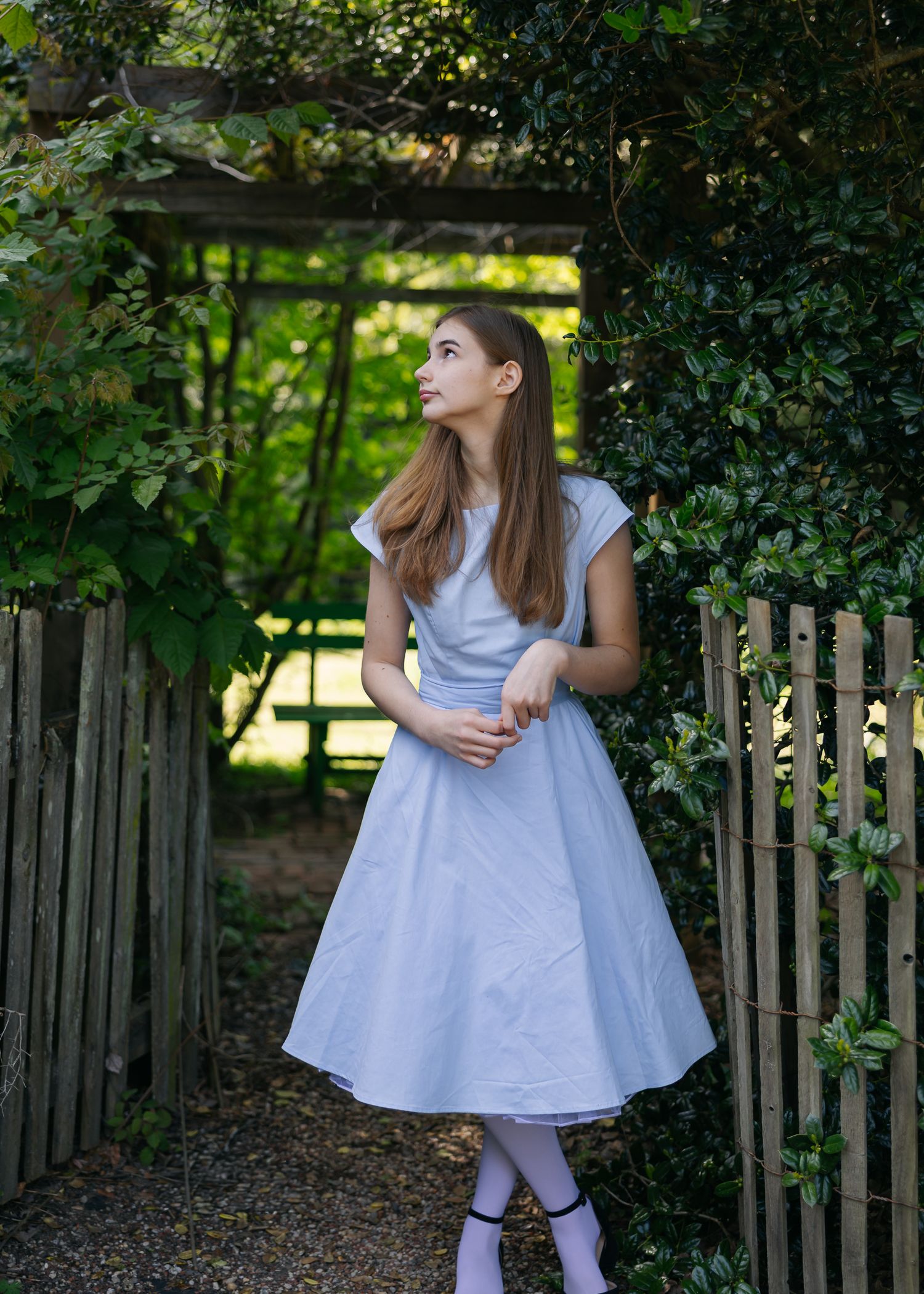 Young woman in a light blue dress standing by a wooden garden gate surrounded by lush green foliage.