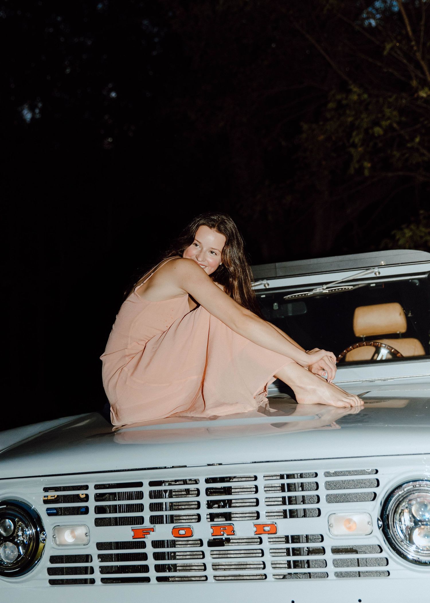 Woman in pink dress sitting on hood of vintage Ford Bronco at night.