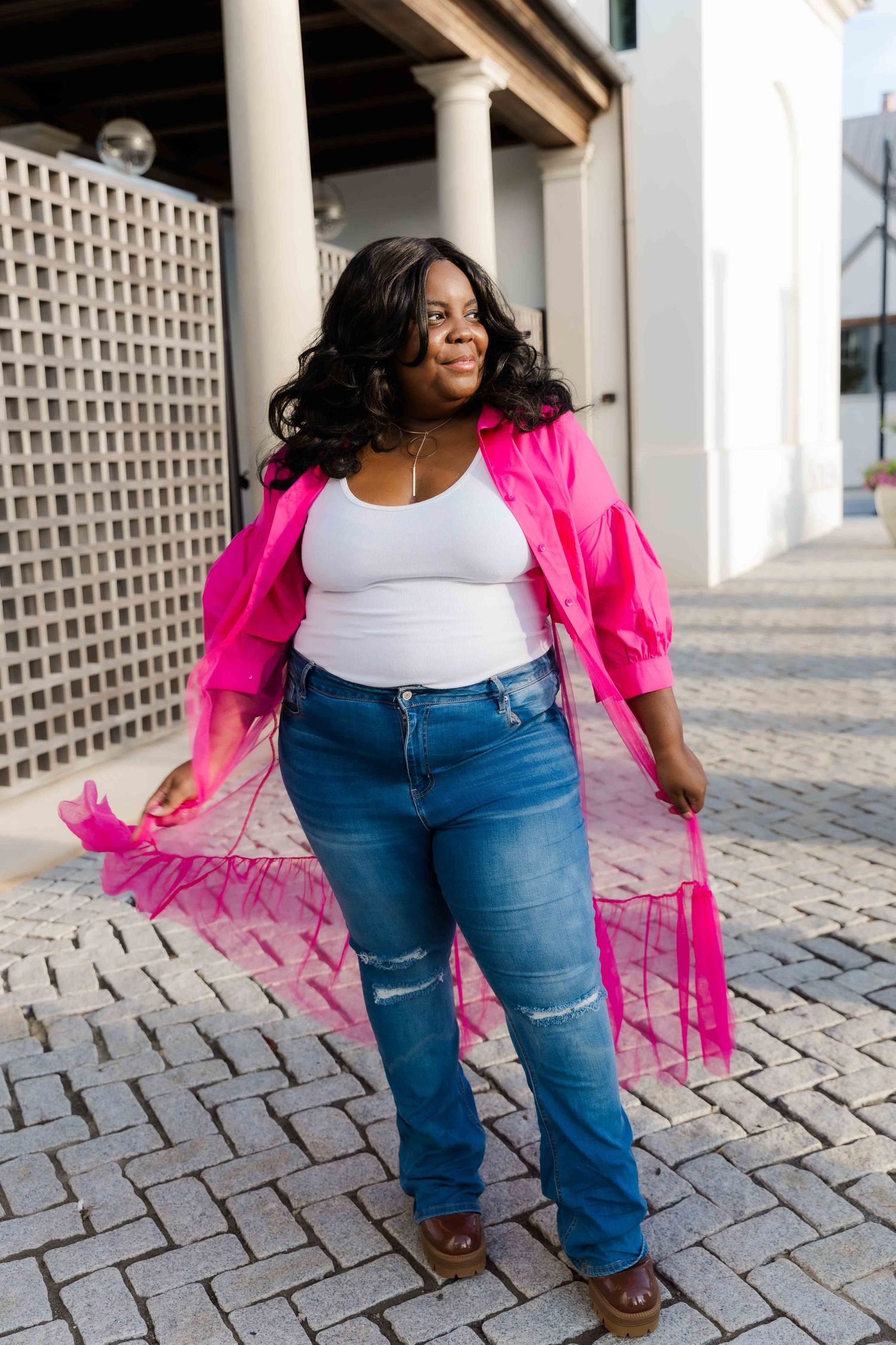 Plus-size woman in pink sheer jacket, white top, and blue ripped jeans posing outdoors on cobblestone walkway.