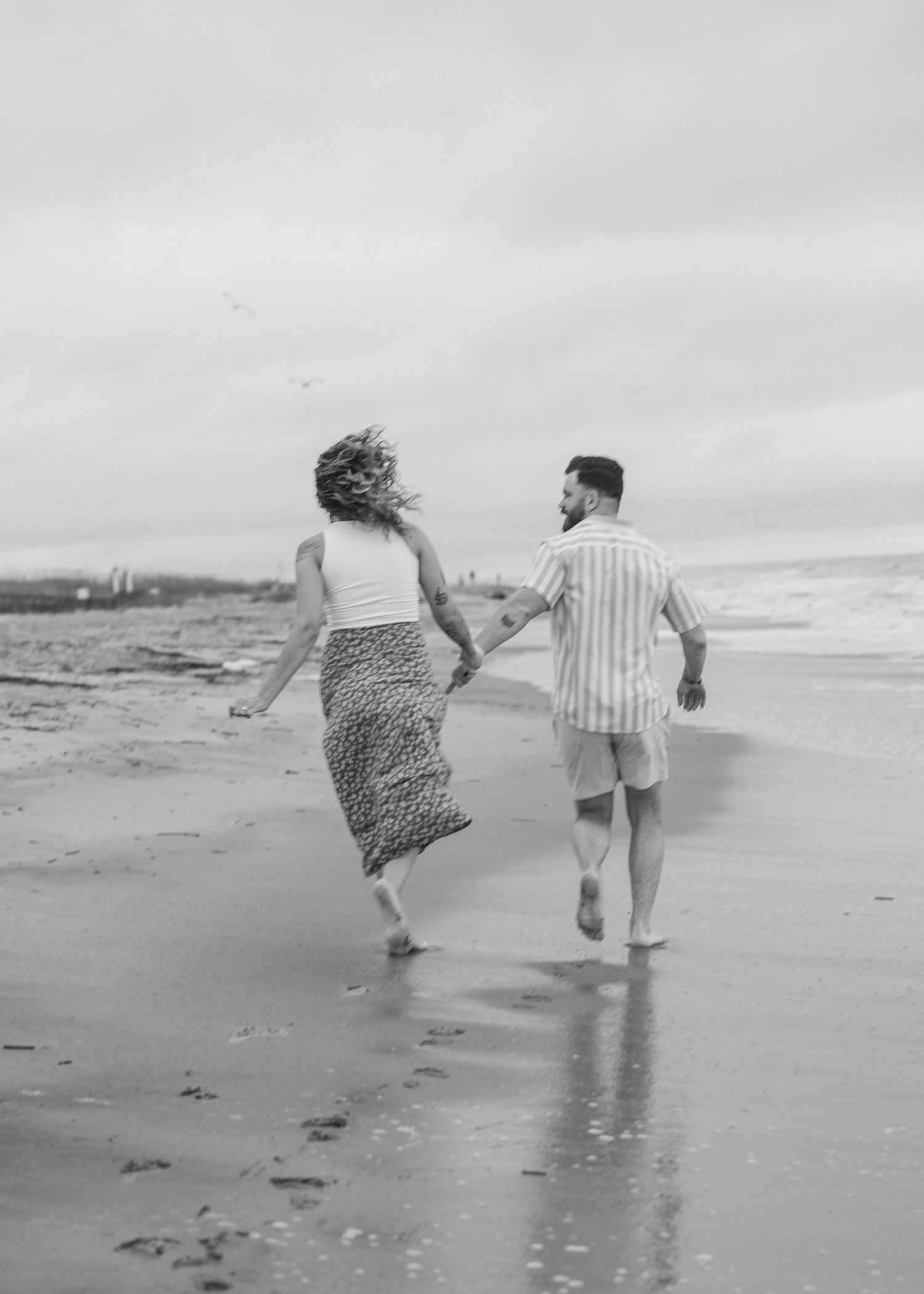 Black and white photos of a couple playfully running through ocean waves on a beach.